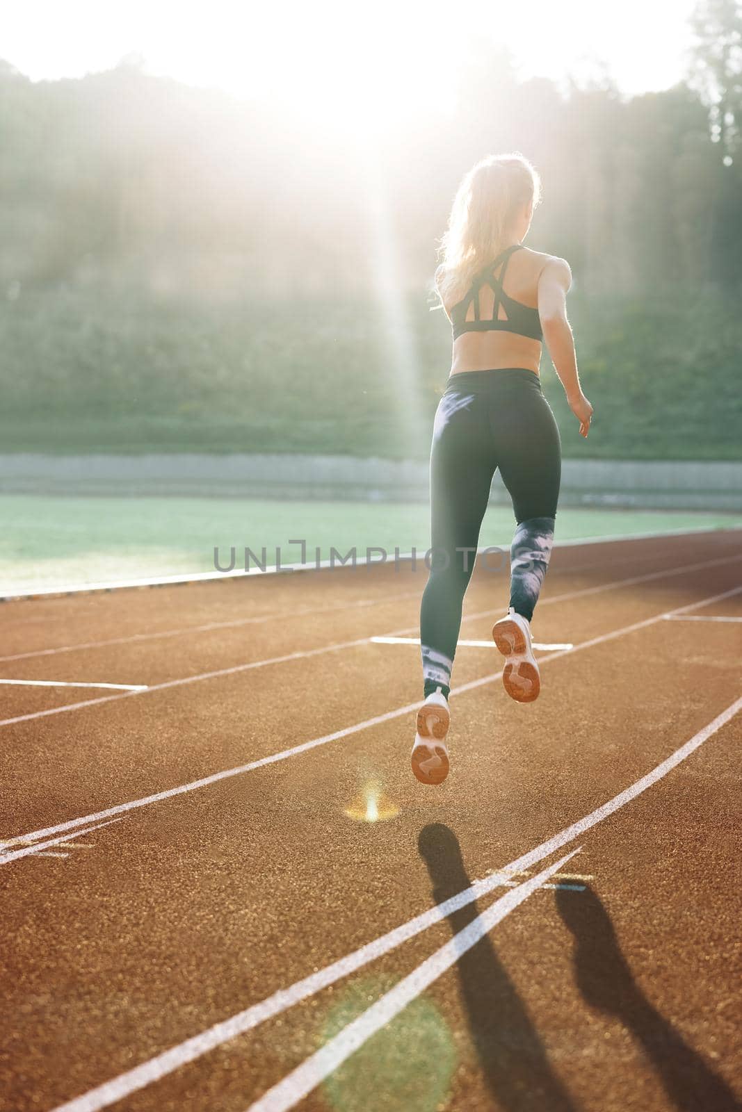 Rear view of athletic woman running on track stadium at summer morning light. Fit woman running on racetrack during training session. Female runner practicing on athletics race track. Sportswoman.