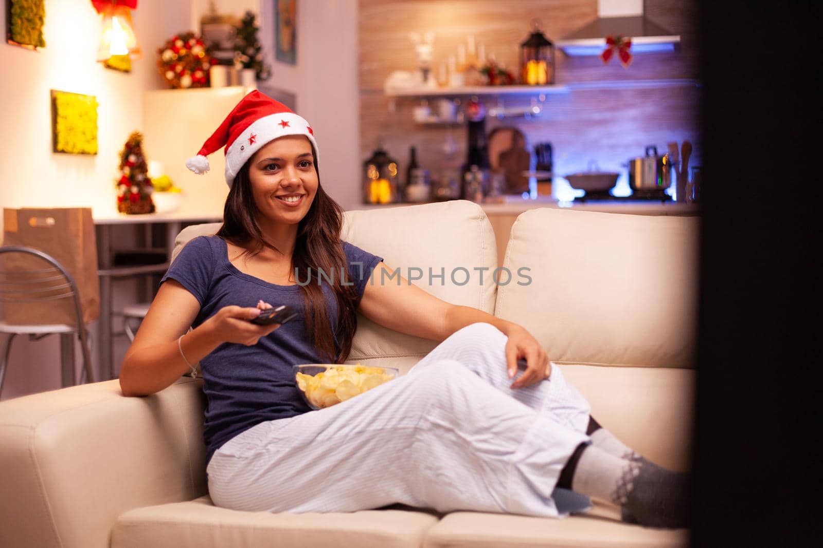 Adult person wearing red santa hat lying on couch watching christmas comedy movie during christmastime in xmas decorated kitchen. Woman enjoying winter season celebrating x-mas holiday