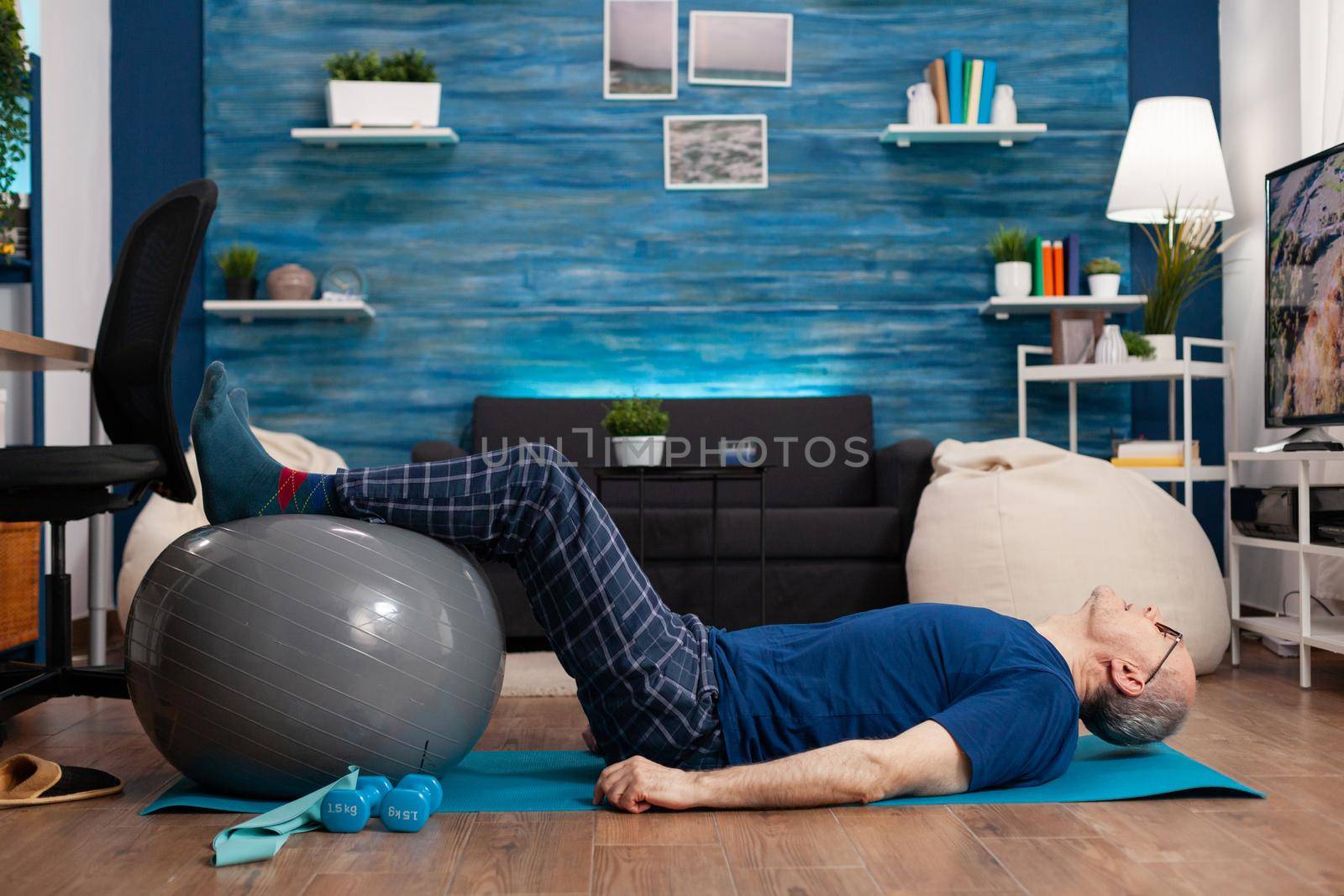 Retired senior sportman sitting on yoga mat practicing warming legs up exercises using swiss ball stretching abdominals muscles. Focused pensioner training body strength resistance in living room