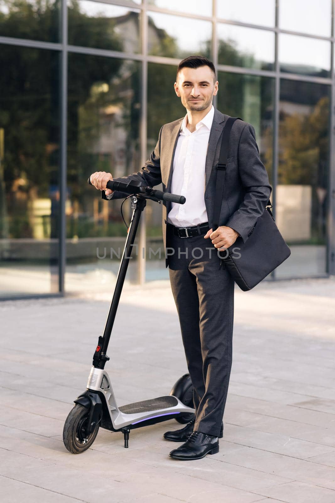 Businessman standing in front of city, holding electric scooter and looking at camera. Modern urban alternative transport. Eco-friendly transportation. Ecology and urban lifestyle by uflypro