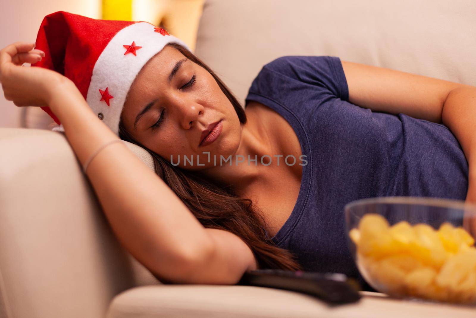Adult person with santa hat sleeping on sofa after watching december entertainment movie on television in xmas decorated kitchen. Woman celebrating christmas holiday enjoying winter season