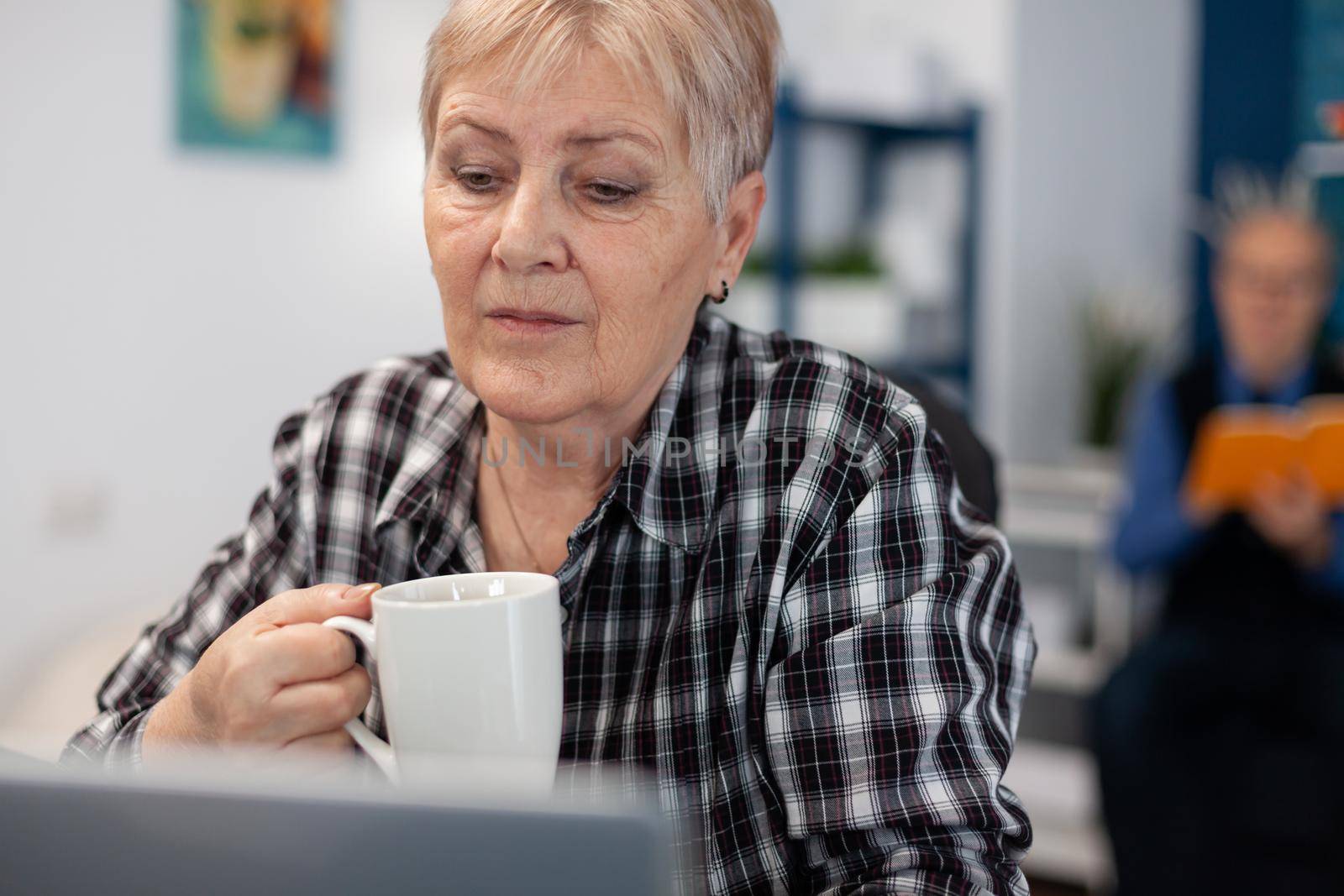 Senior pensioner with cup of coffee in hands reading morning news. Elderly woman in home living room using moder technoloy laptop for communication sitting at desk indoors.
