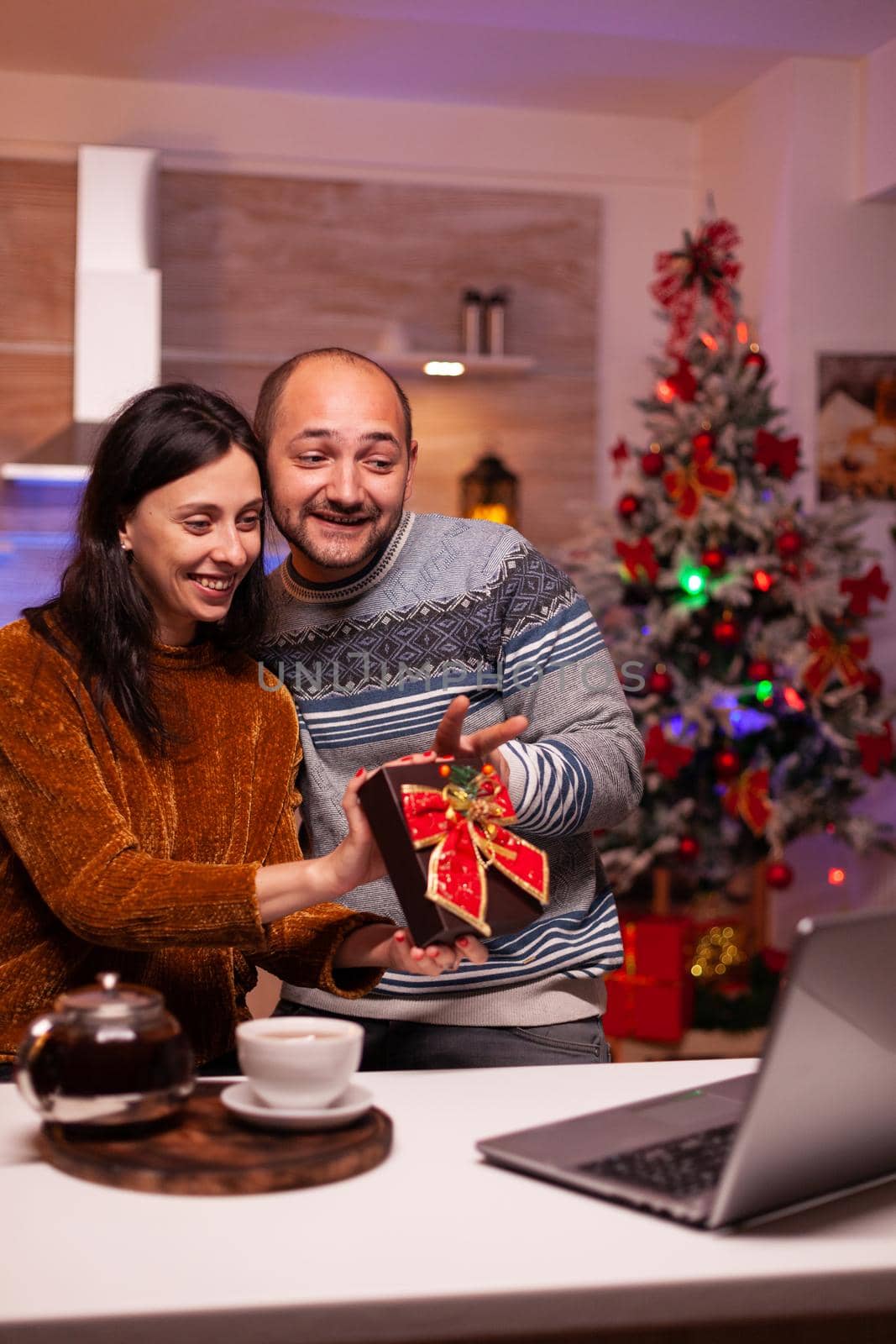 Happy couple showing present to remote friends during online videocall meeting on laptop computer. Joyful smiling family enjoying winter season celebrating christmas holiday in xmas decorated kitchen