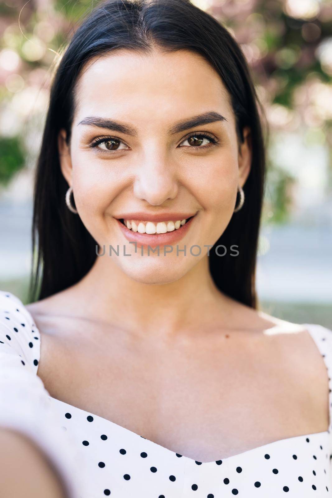Vertical format photo of beautiful joyful young Caucasian woman in white dress posing to smartphone camera and taking selfie photo. Pretty stylish female tourist making photos with mobile phone by uflypro