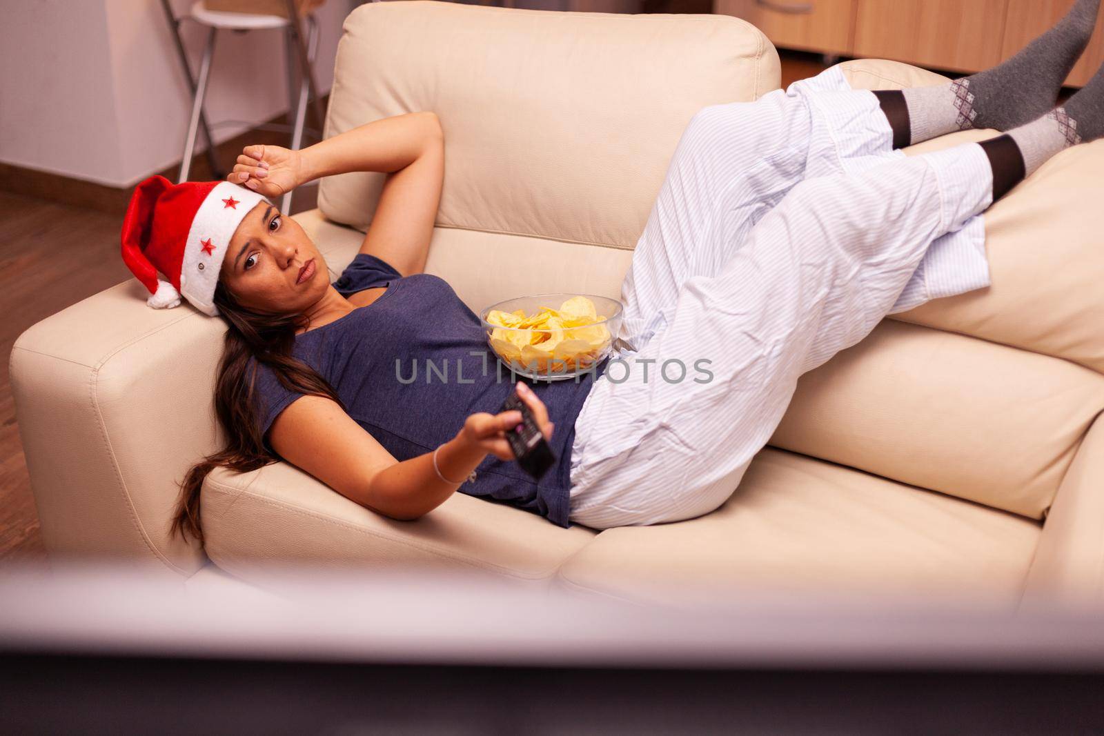 Relaxed woman resting on couch watching christmas movie series on television celebrating christmastime in x-mas decorated kitchen. Girl wearing santa hat enjoying winter season at home