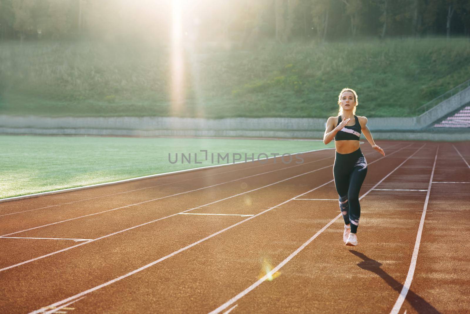 Front view of running caucasian woman in black sportswear doing cardio workout on sports track of stadium. Adult female athlete jogging, training before marathon competition. Health. Sportswoman. by uflypro