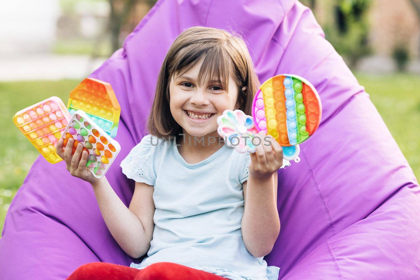 Portrait little girl with a modern popit toy. Colorful and bright pop it toy. Simple dimple. The child is sitting on a easy chair with colorful trendy antistress sensory toy pop it and simple dimple.