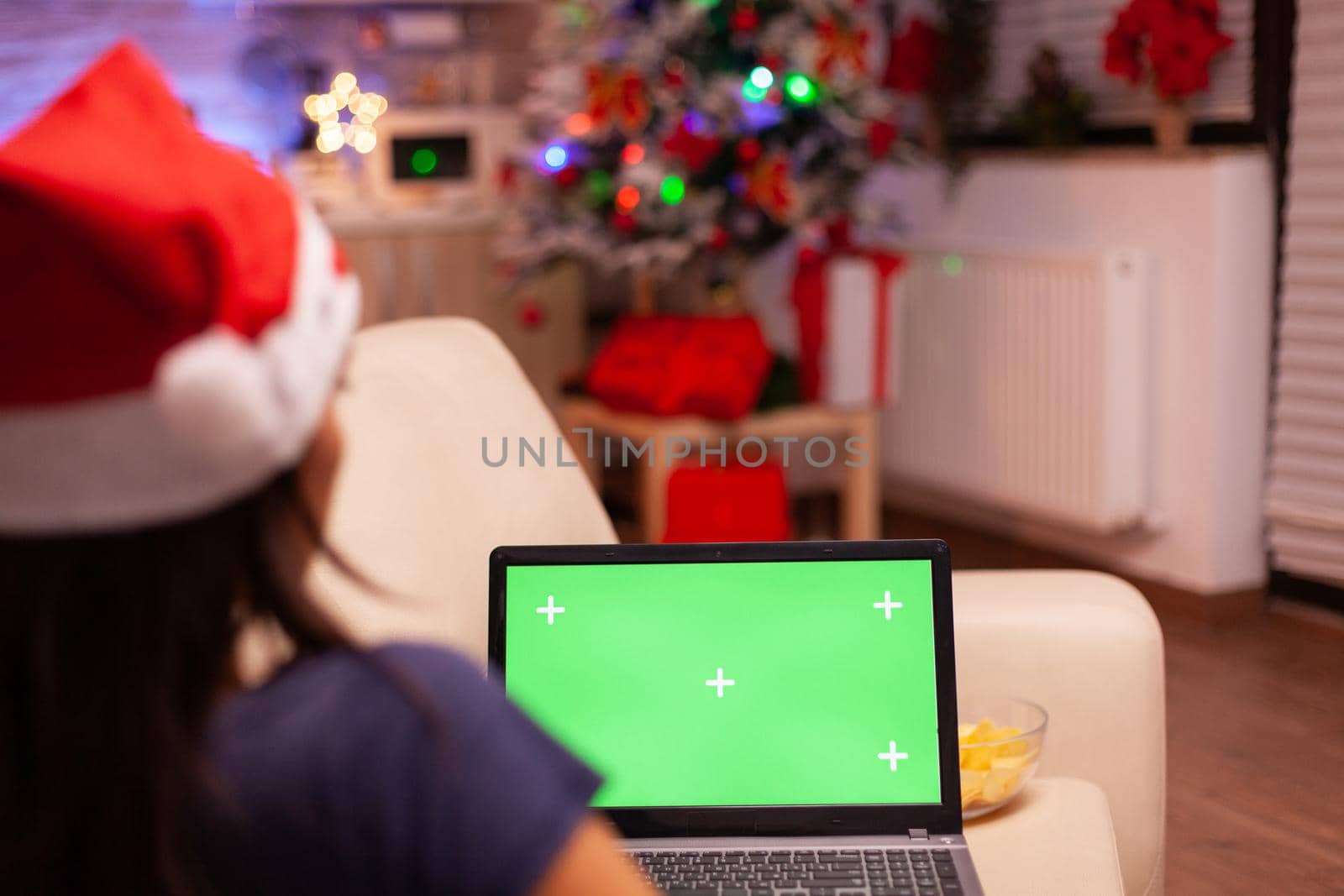 Woman sitting on couch in xmas decorated kitchen holding green screen mock up chroma key laptop computer with isolated display. Adult person enjoying christmas holiday celebrating winter season