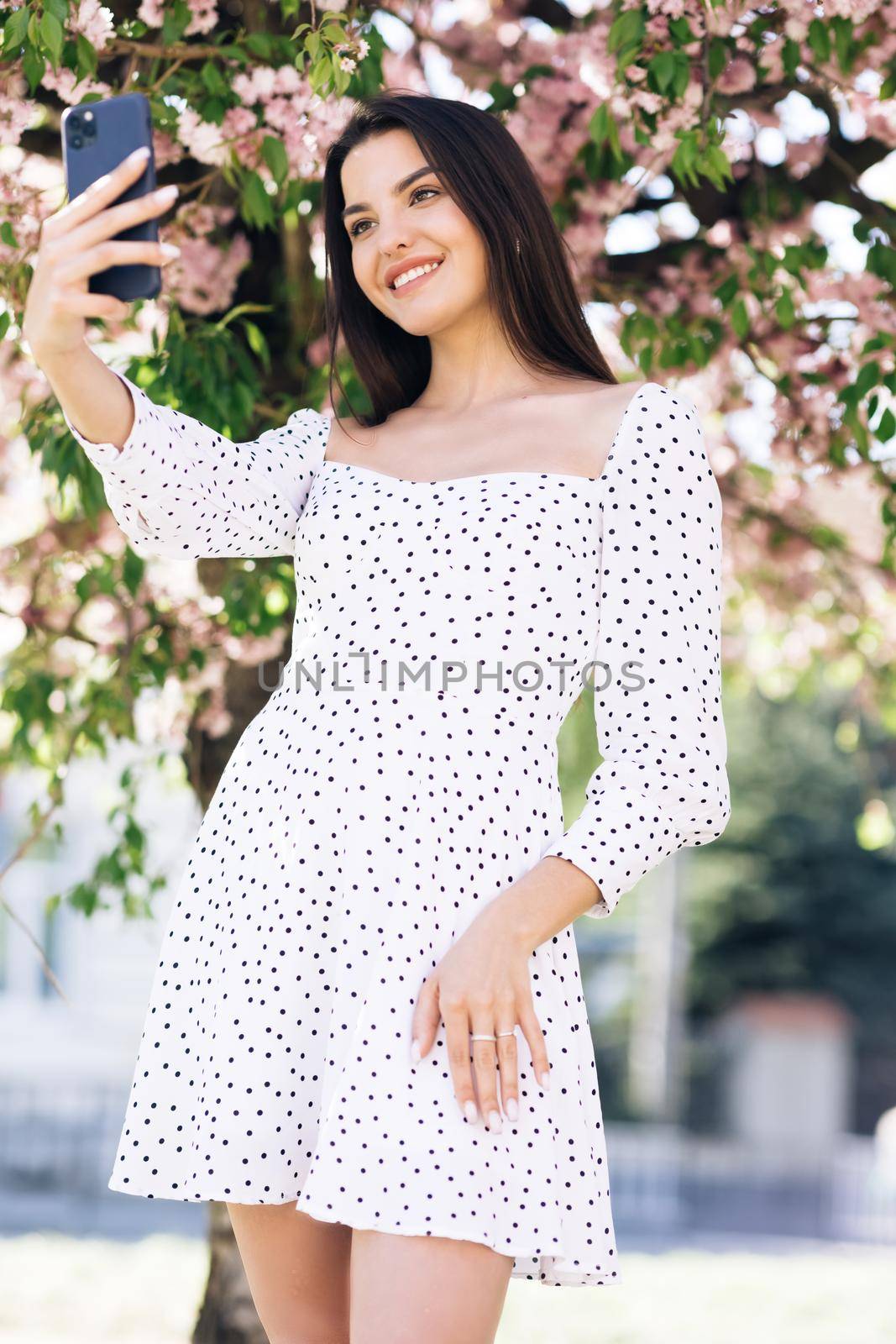 Young smiling woman in summer white dress taking selfie self portrait photos on smartphone. Model posing on park sakura trees background. Female showing positive face emotions. by uflypro