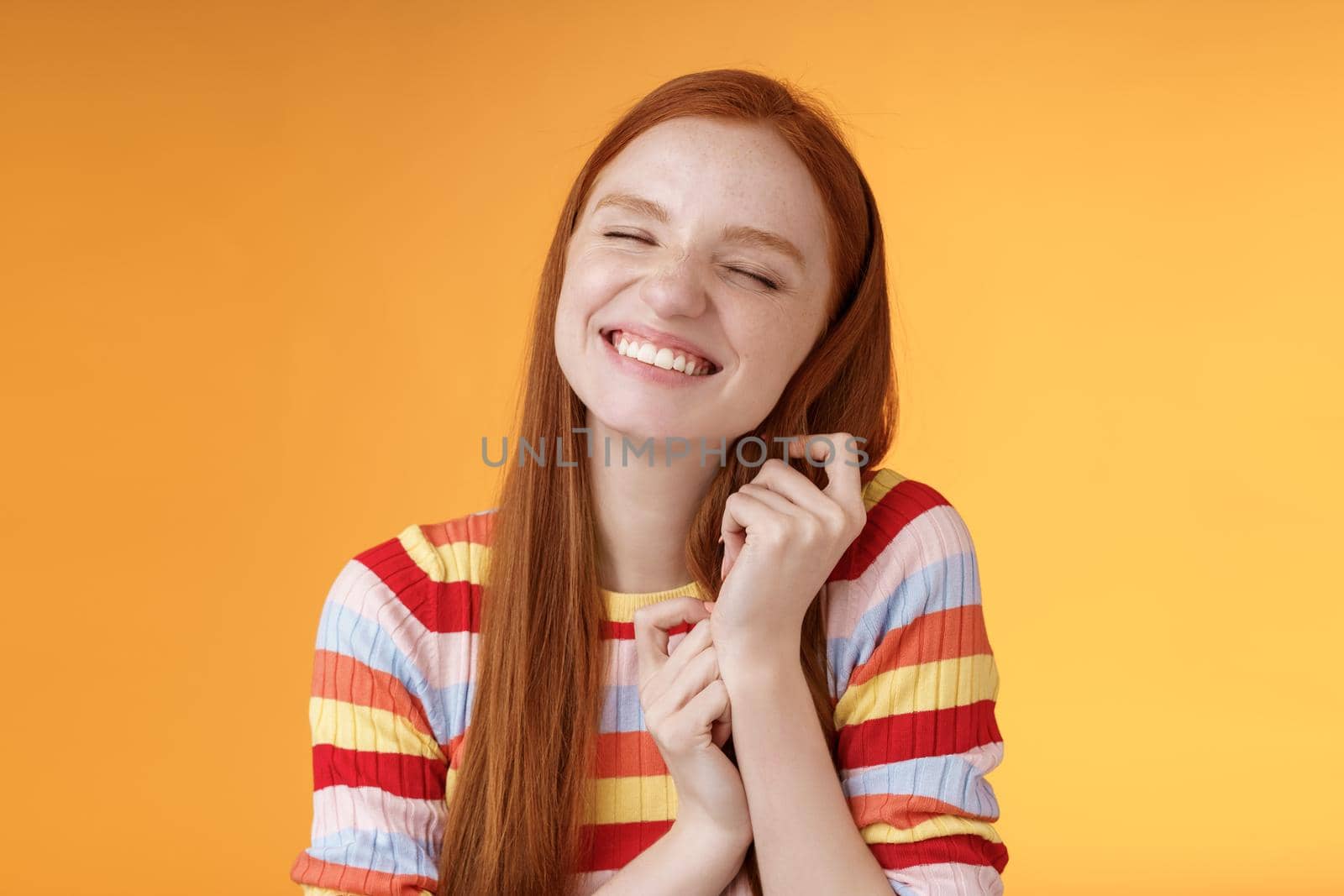 Happy dreamy romantic young tender ginger girl fantasizing creating love story imagination smiling broadly delighted close eyes touching hair strands recalling nice memory, standing orange background.