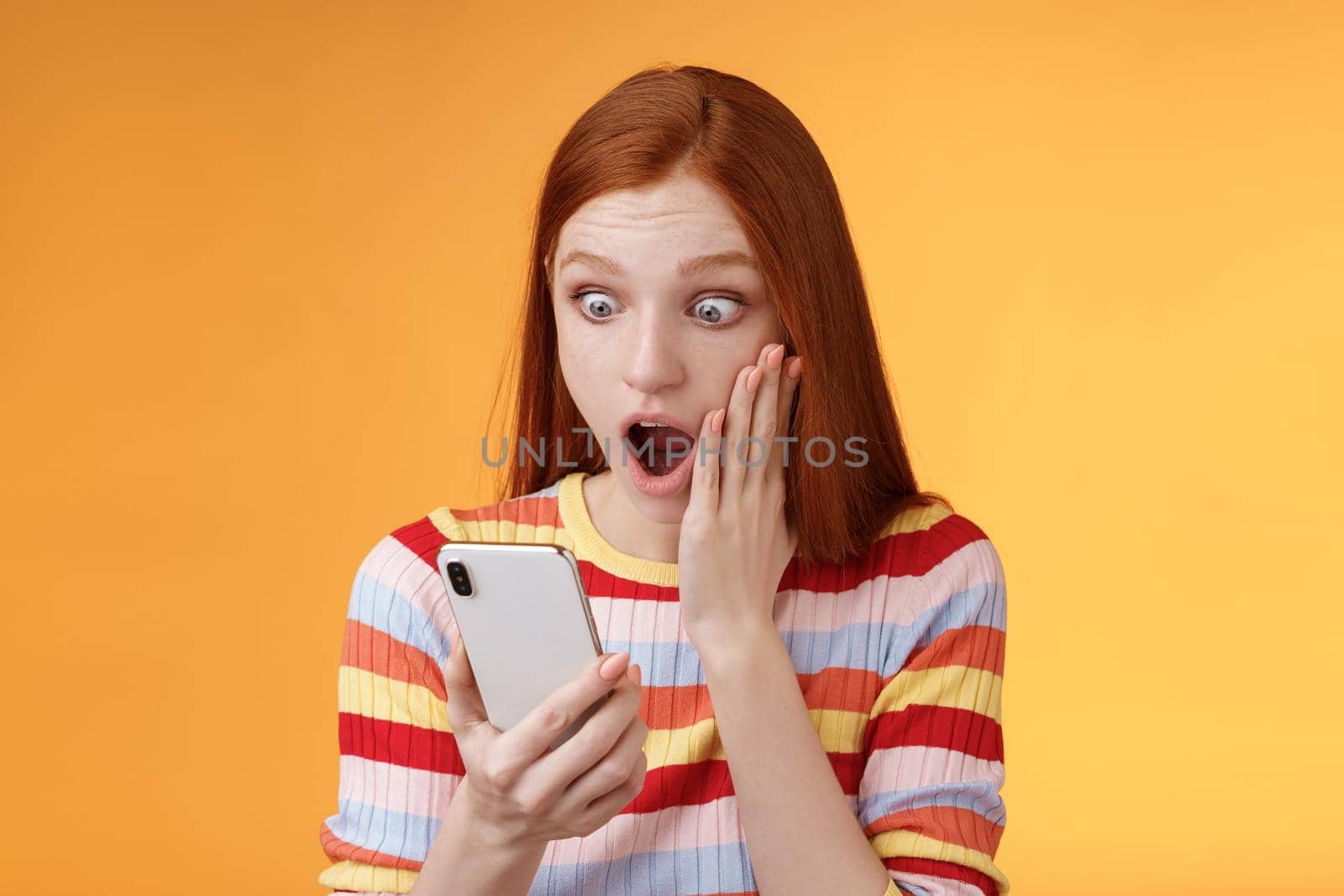 Omg what heck. Portrait shocked concerned young redhead sensitive impressed redhead woman stare smartphone display touch cheek drop jaw stunned surprised standing orange background hold phone.