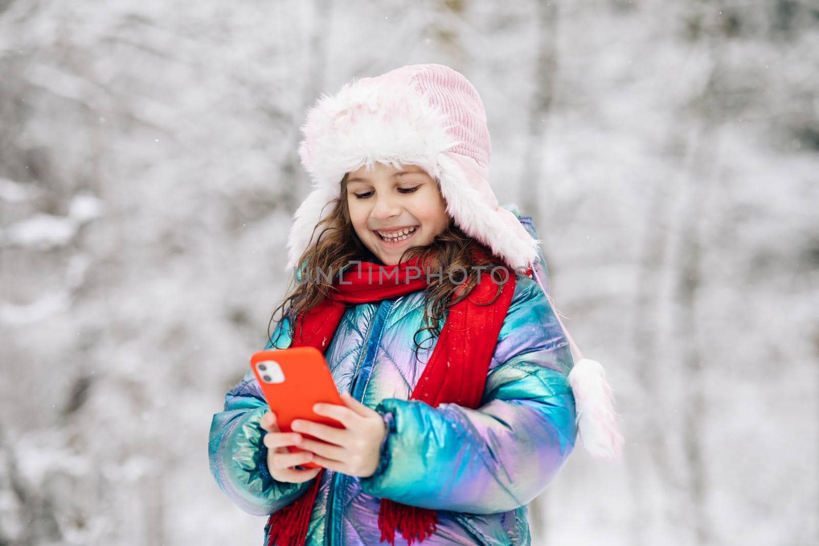 Happy little child use smartphone funny face mask app look at cell screen smiles at something funny. Holidays and celebrations concept. Outside. The dead of winter, cold weather, frost, snow