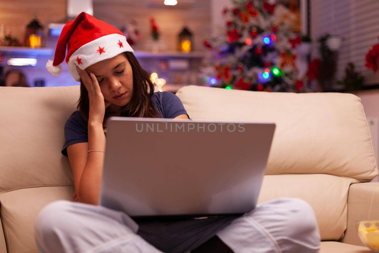 Tired exhausted woman sleeping on sofa in xmas decorated kitchen while searching business email on laptop computer. Caucasian female celebrating christmas holiday enjoying winter season