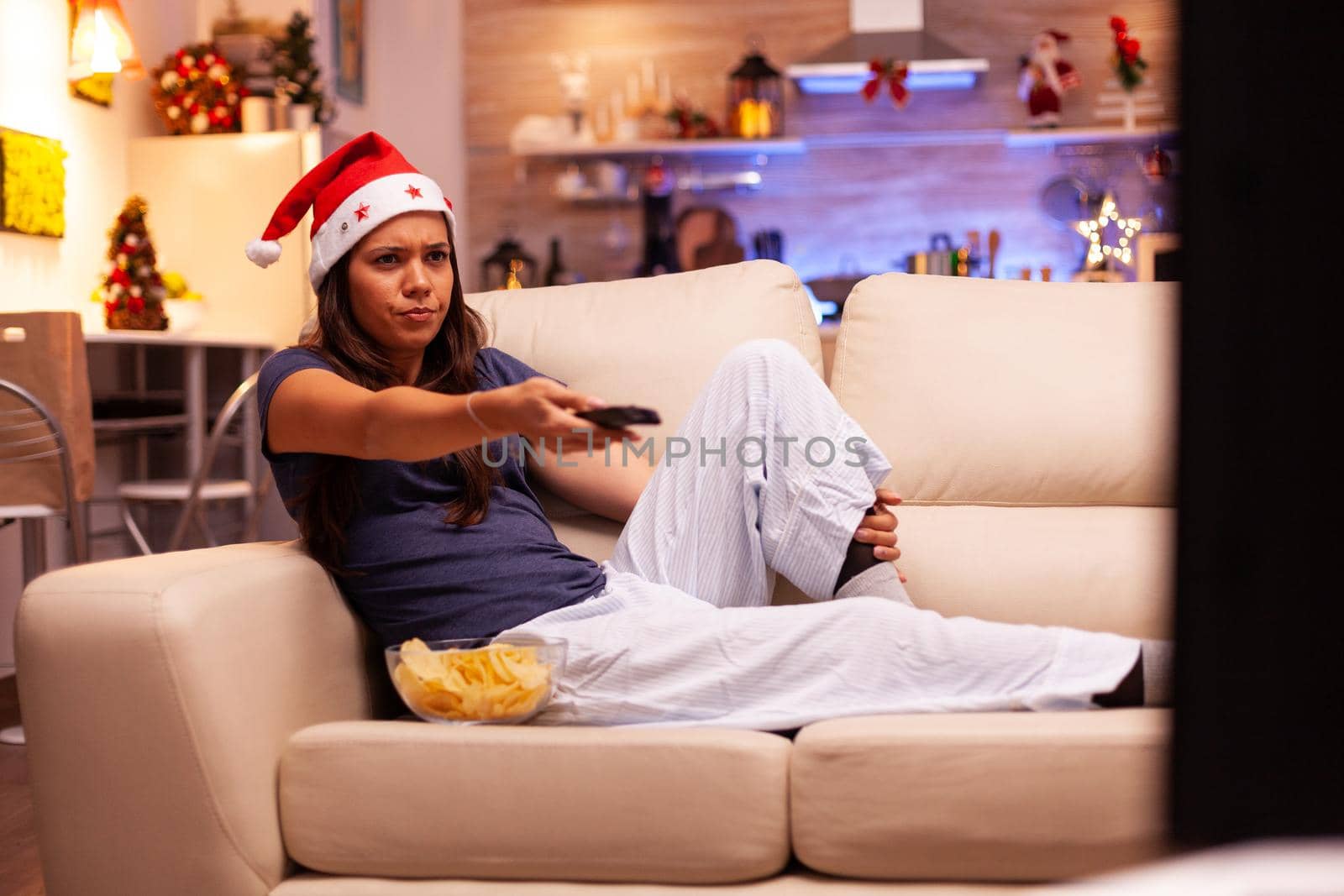 Caucasian female resting on couch watching entertainment movie series on television using remote control enjoying winter season in xmas decorated kitchen. Woman celebrating christmas holiday