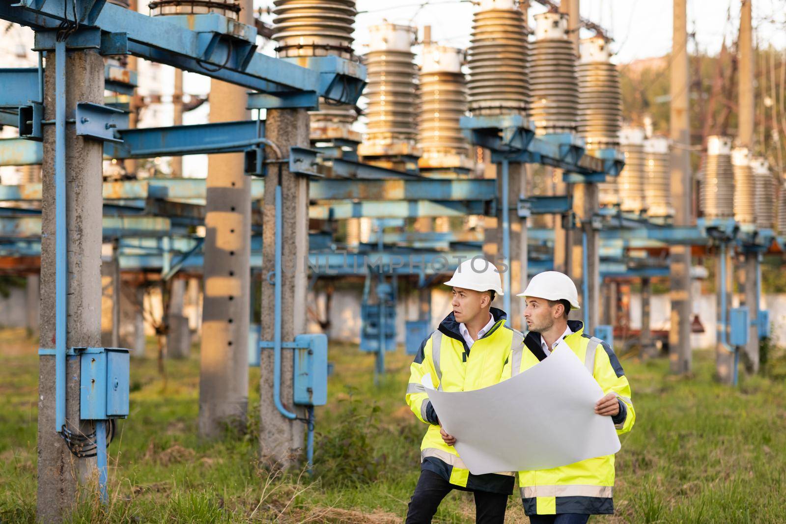 Engineers in special clothing discuss a drawing on paper near high-voltage power line. Engineers men in warm clothes use construction plan discuss work walking against power electric station by uflypro