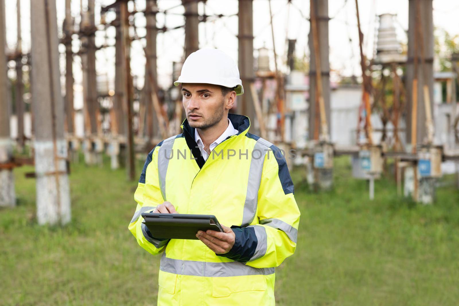Male engineer in uniform and helmet standing outdoor and tapping on tablet. Man standing near high voltage substation and typing on device.