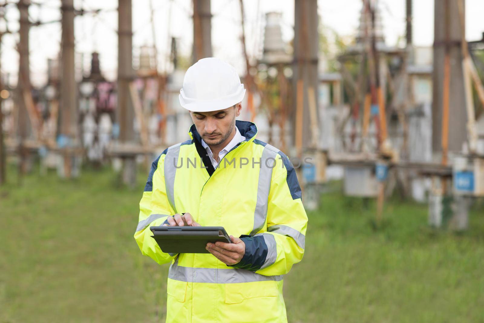 Man standing near high voltage substation and typing on device. Male engineer in uniform and helmet standing outdoor and tapping on tablet by uflypro