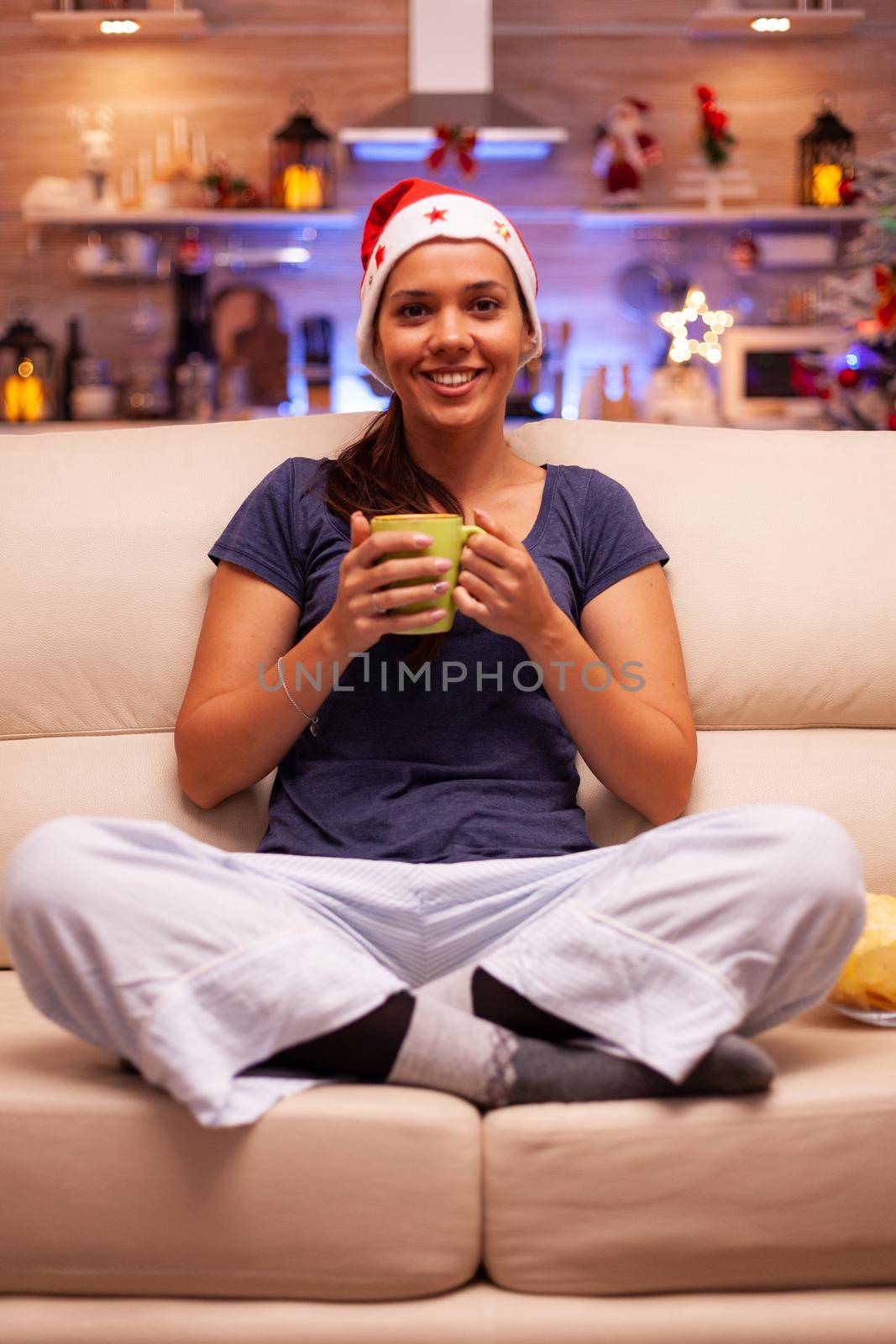 Portrait of smiling woman wearing red santa hat sitting in lotus position on couch enjoying winter holiday. Girl in comfy clothes celebrating christmas season in xmas decorated kitchen