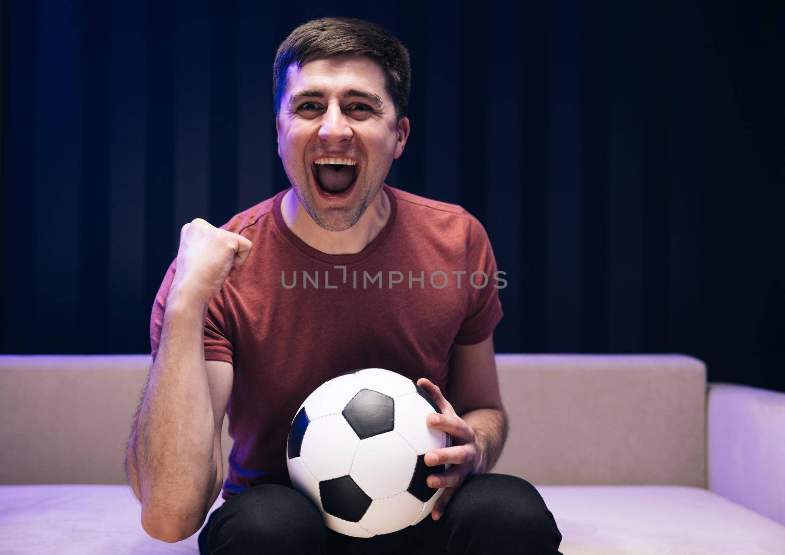 People emotions sport leisure lifestyle concept. Young fun guy 30s football fan cheer up support favorite team hold ball in red t-shirt in dark living room.