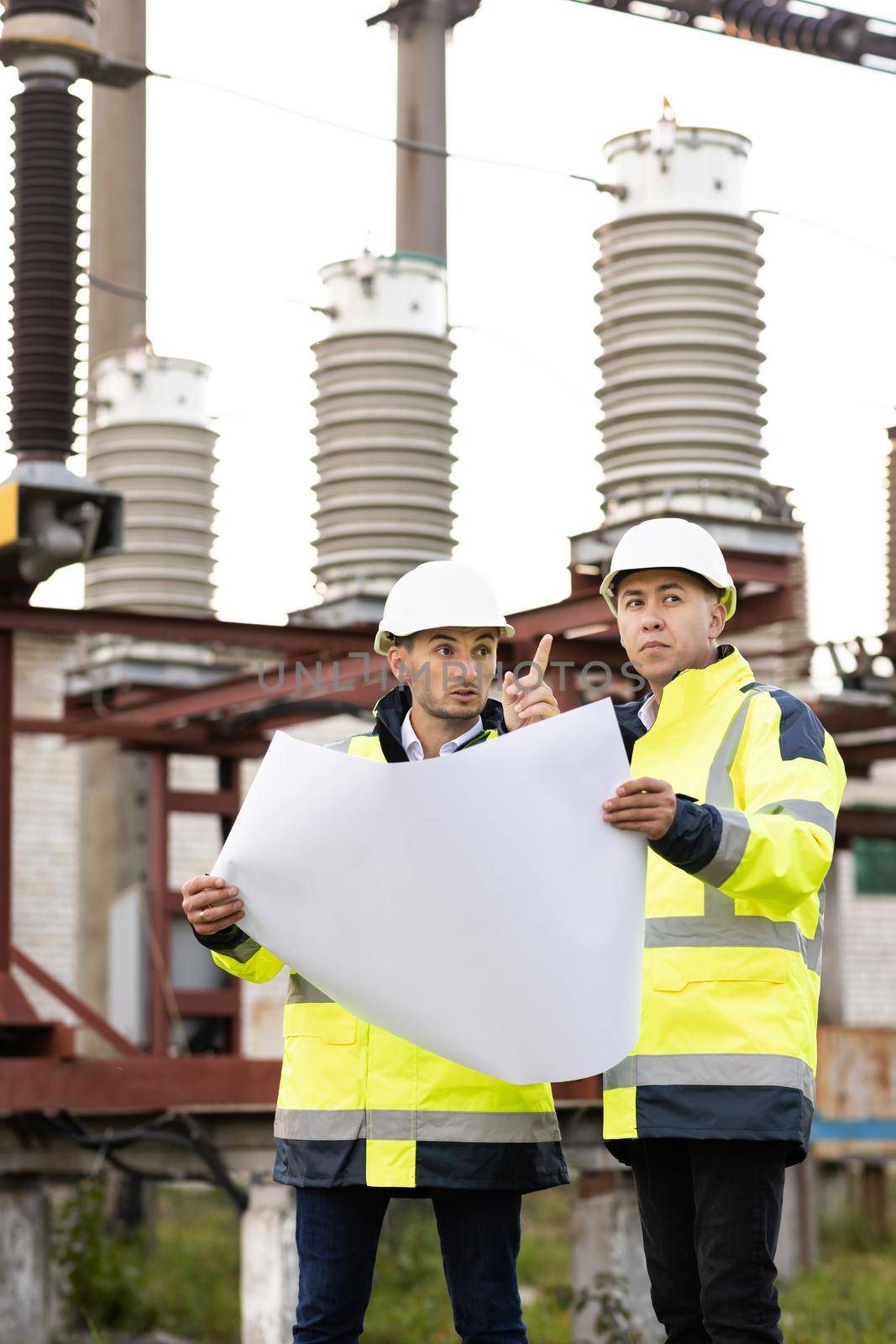 Engineers near high-voltage powerline working with a construction plan. Two engineers in special clothing discuss a drawing on paper against the background of a high-voltage power line.