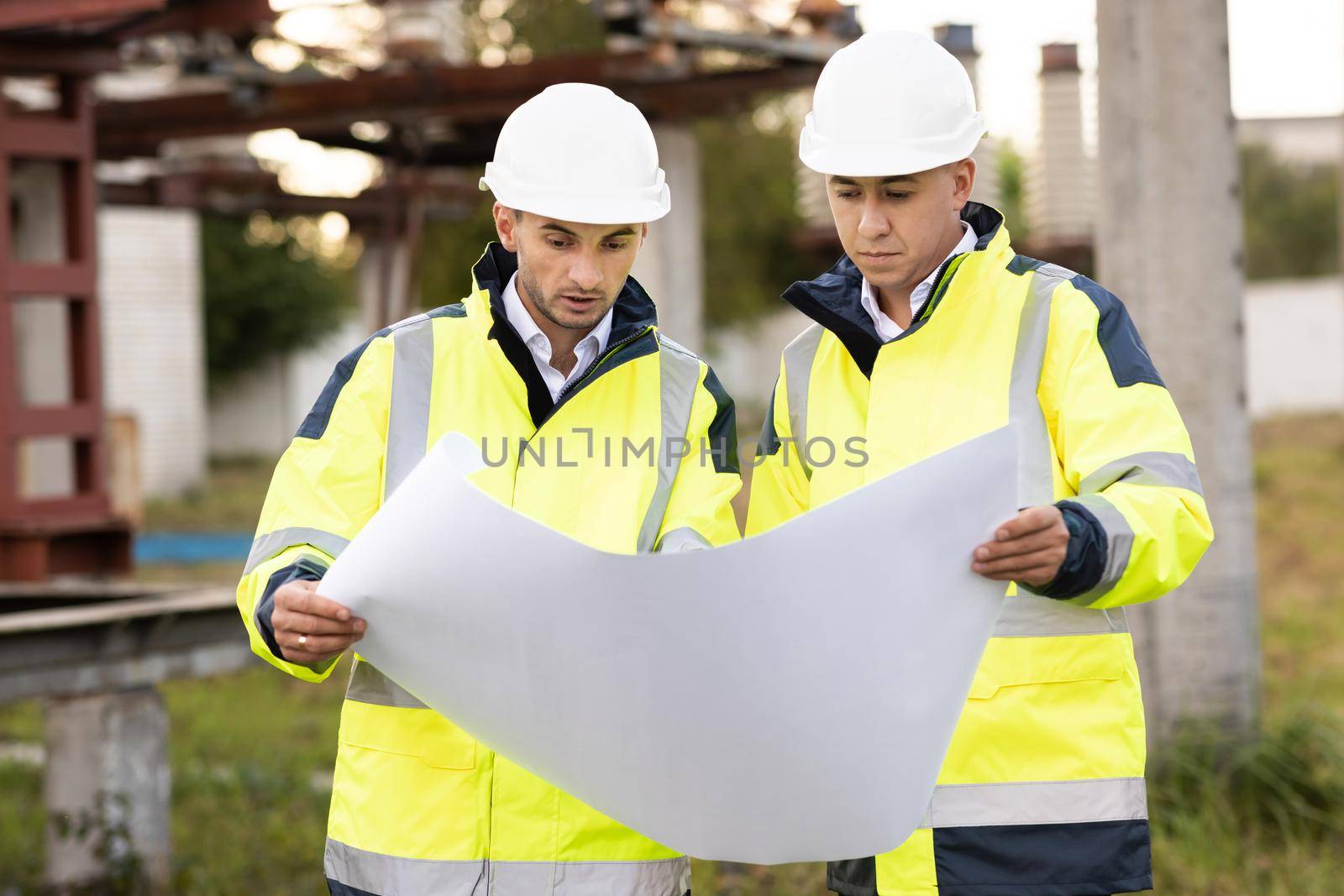 Two engineers are talking in front of electrical transmission lines, working on renewable energy development. Engineers in special clothing discuss a drawing on paper near high-voltage power line by uflypro