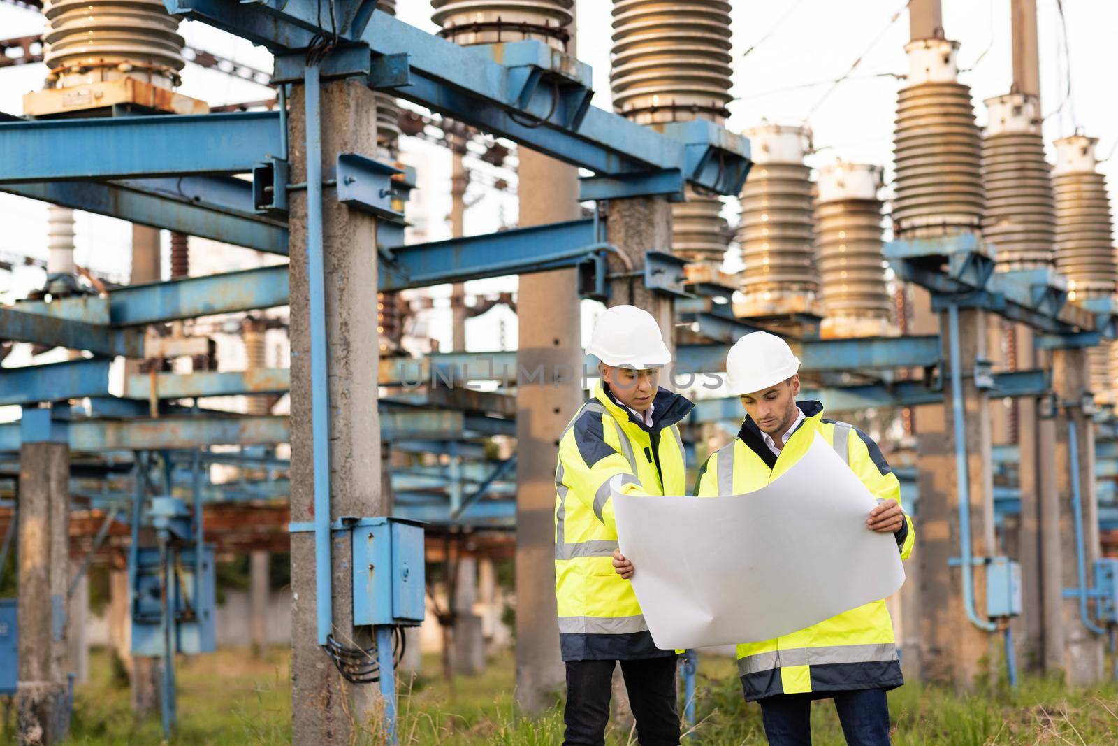 Engineers men in warm clothes use construction plan discuss work walking against power electric station. Engineers in special clothing discuss a drawing on paper near high-voltage power line by uflypro