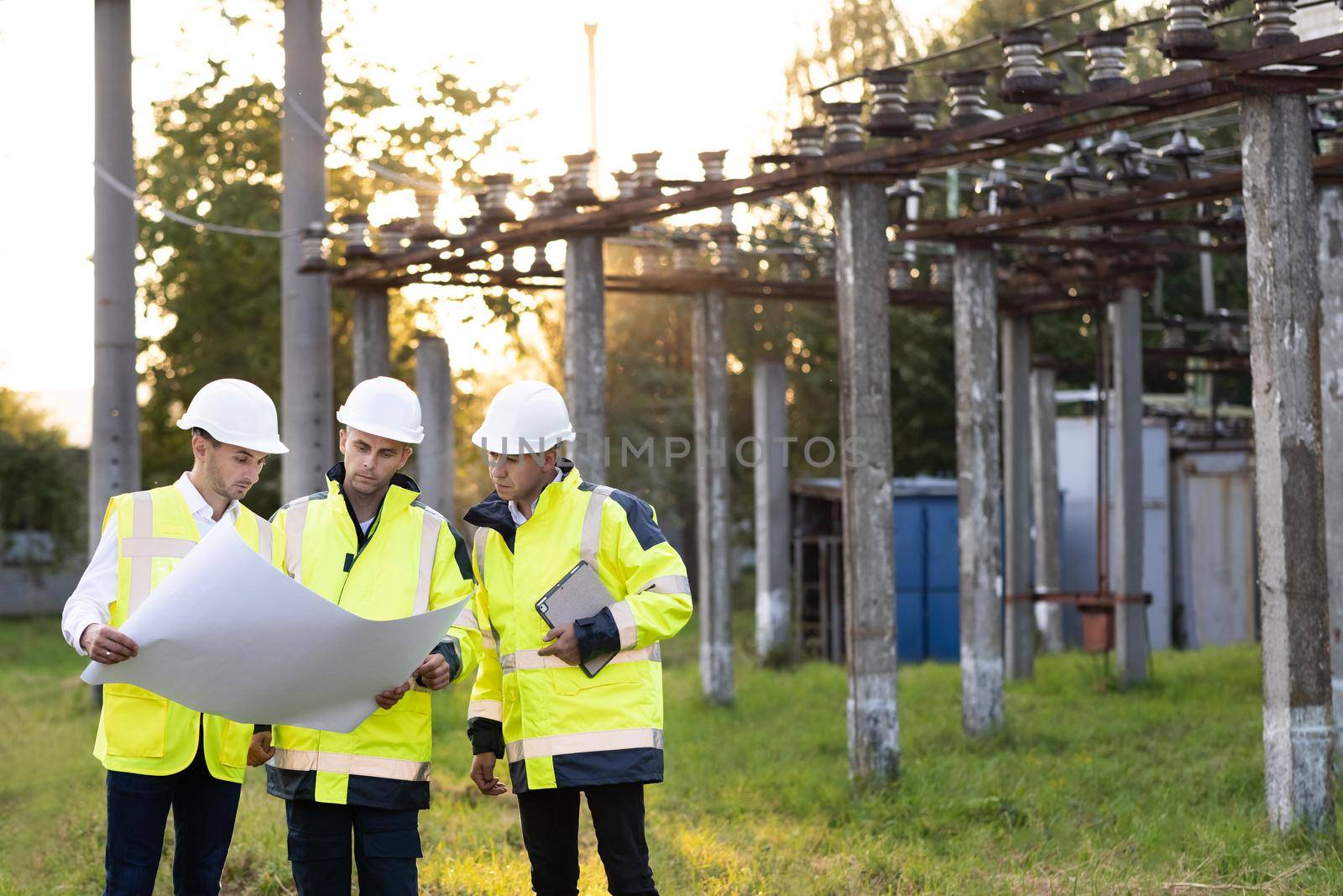 Power specialists are planning a new project outdoors. Three engineers walk near power lines in the high voltage power station. Electric industry, electrical energy production concept.