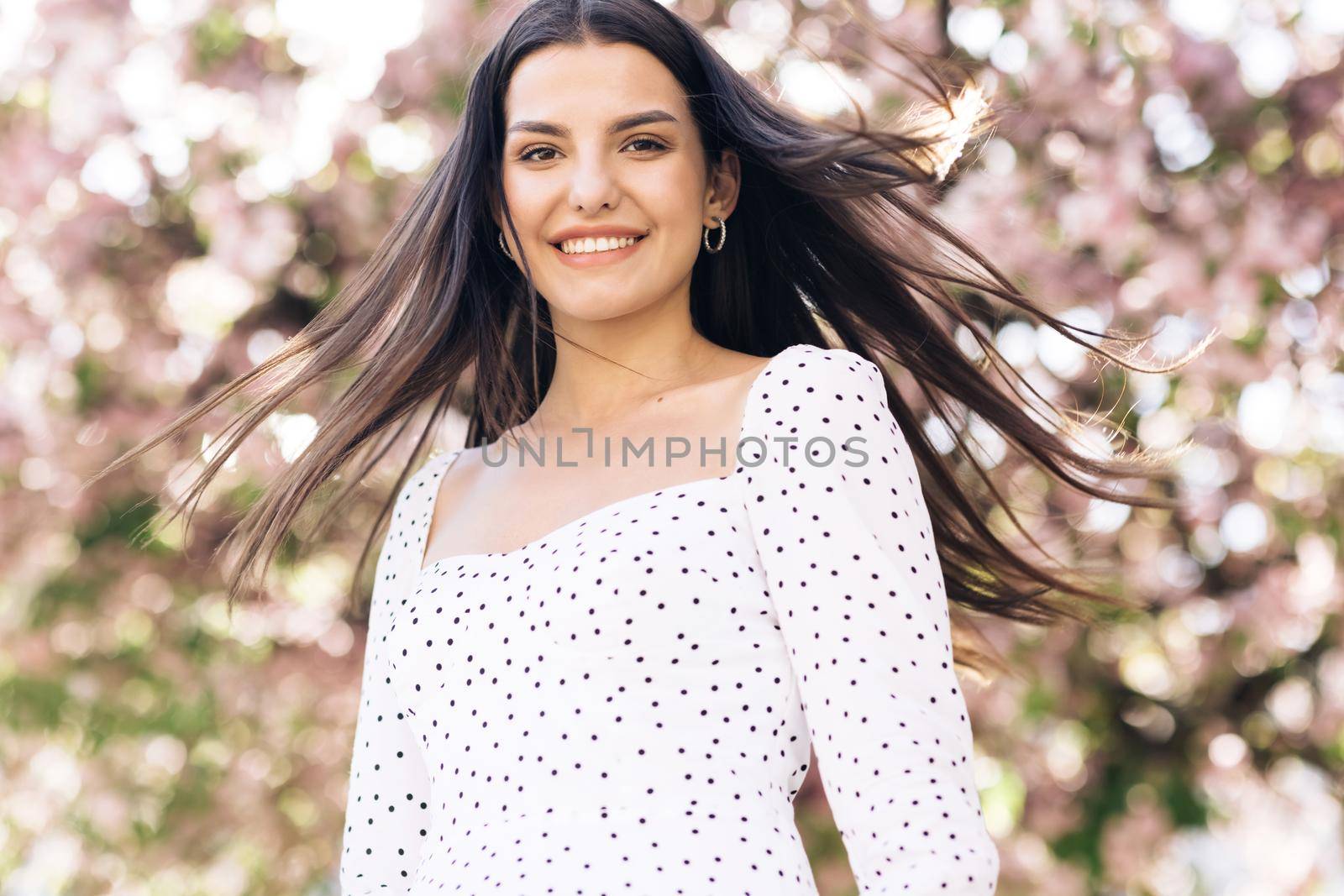 Portrait attractive sunshine young smiling woman with long hair look at camera smile portrait happy summer face sunset beautiful lady outdoor. Spring blossom of sakura tree in city park by uflypro