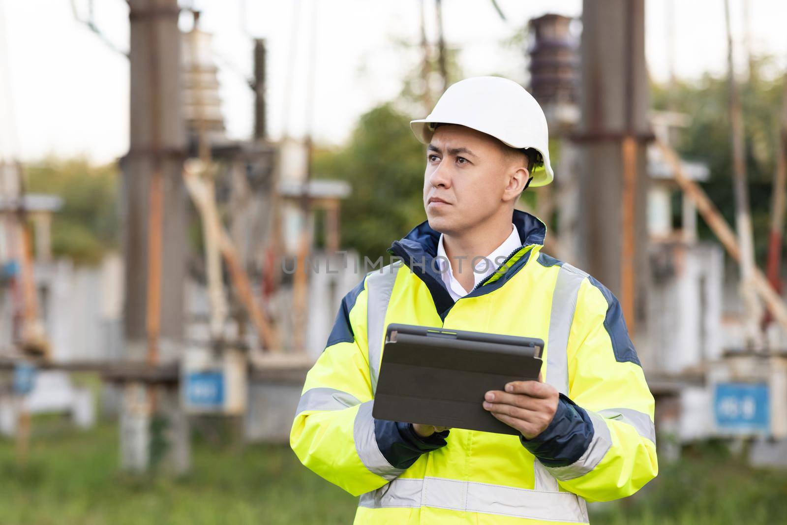 Electrical engineer with high voltage electricity pylon. An energy engineer man in special clothes inspects a power line using data from electric sensors on a digital tablet computer by uflypro