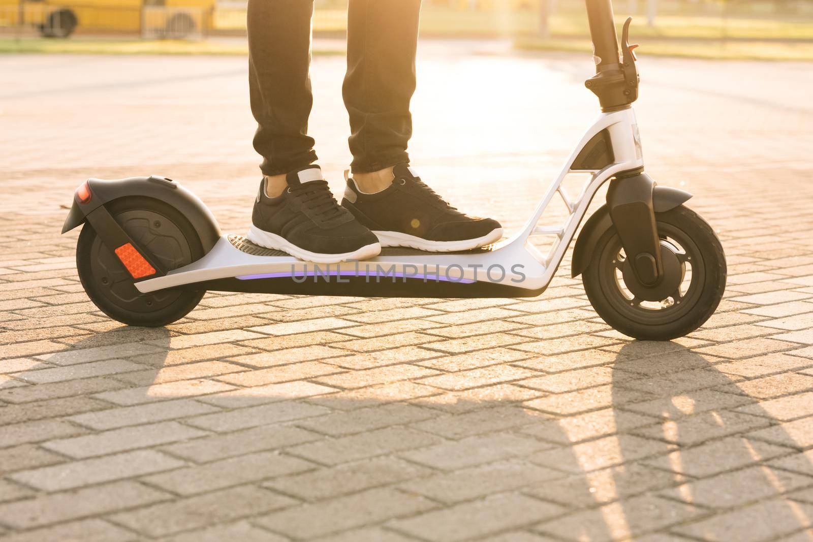 Shot on legs young hipster man in black sneakers ride on electric mobile scooter at sunset. Man hipster riding an electric scooter. Eco-friendly transportation. Fast speed driving electric transport by uflypro