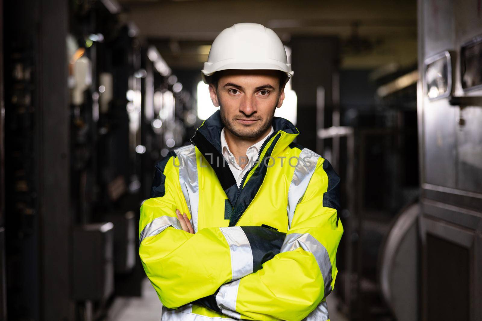 Young factory engineer or worker wearing safety vest and hard hat crossing arms at electrical control room. Industry and engineering concept by uflypro