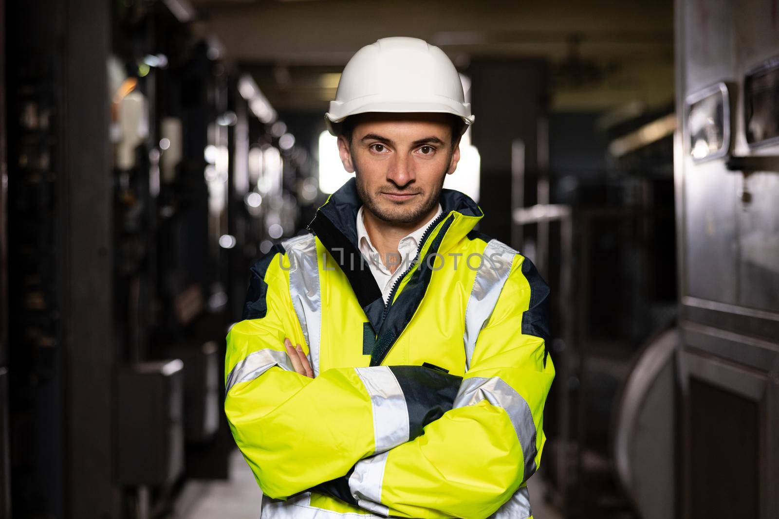 Young factory engineer or worker wearing safety vest and hard hat crossing arms at electrical control room. Industry and engineering concept.