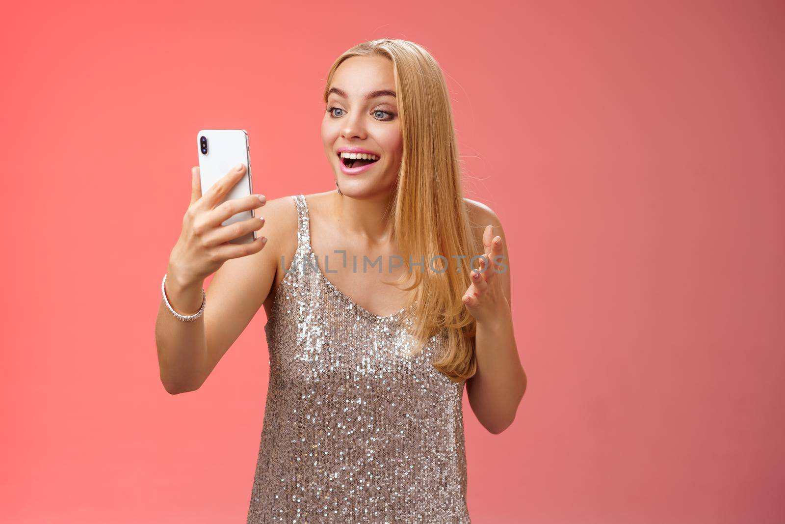 Charming elegant nice blond girl in silver dress talking video call speaking looking smartphone display amused surprised smiling happily have conversation sibling showing prom outfit.