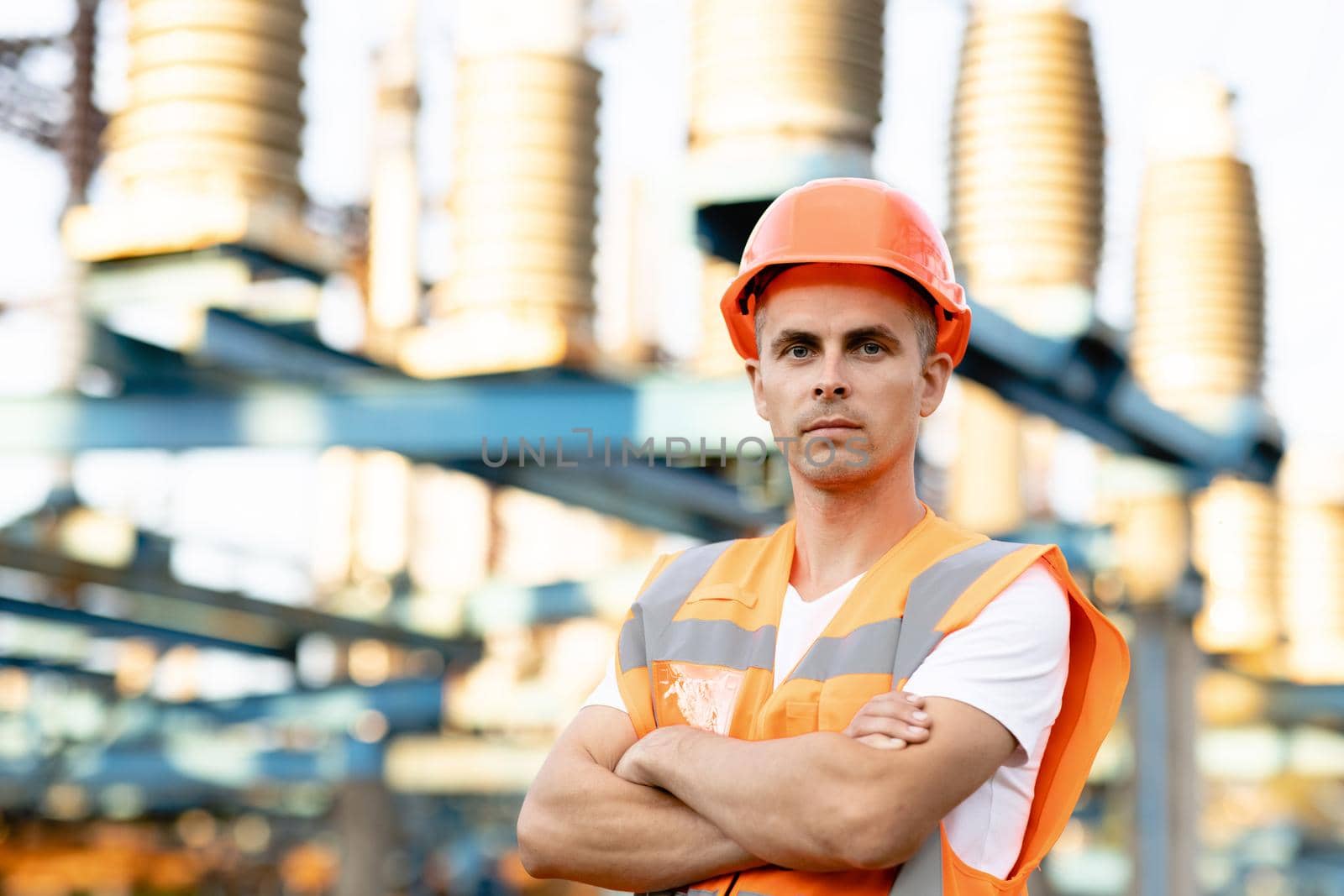 Portrait of engineer worker in uniform and helmet standing near high voltage substation with tall pylons and voltage distribution cables.