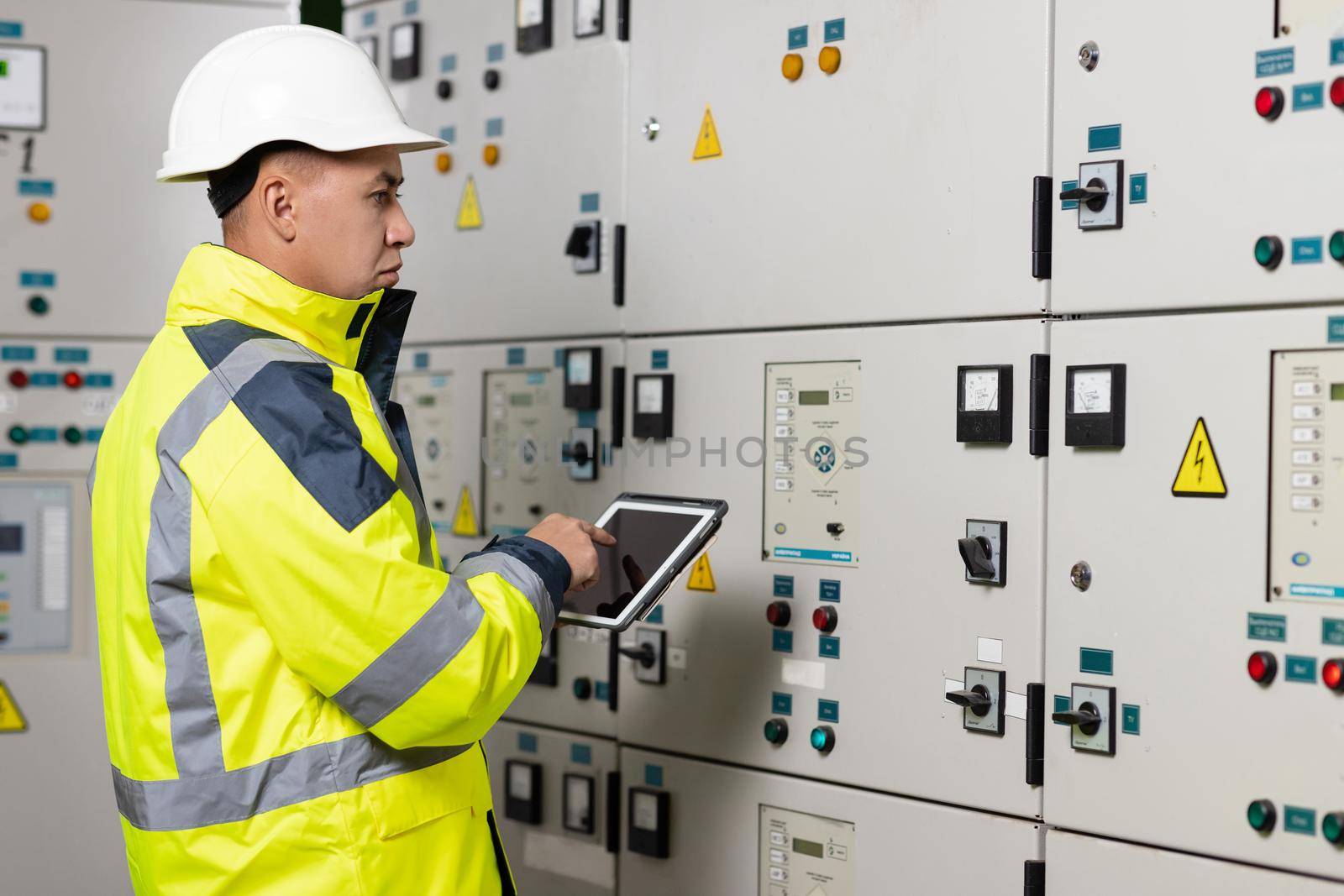 Asian man electrical engineer hold tablet monitoring electrical system in control room. Technician inspect to control panel screen system for generate electricity of factory in manufacture industrial by uflypro