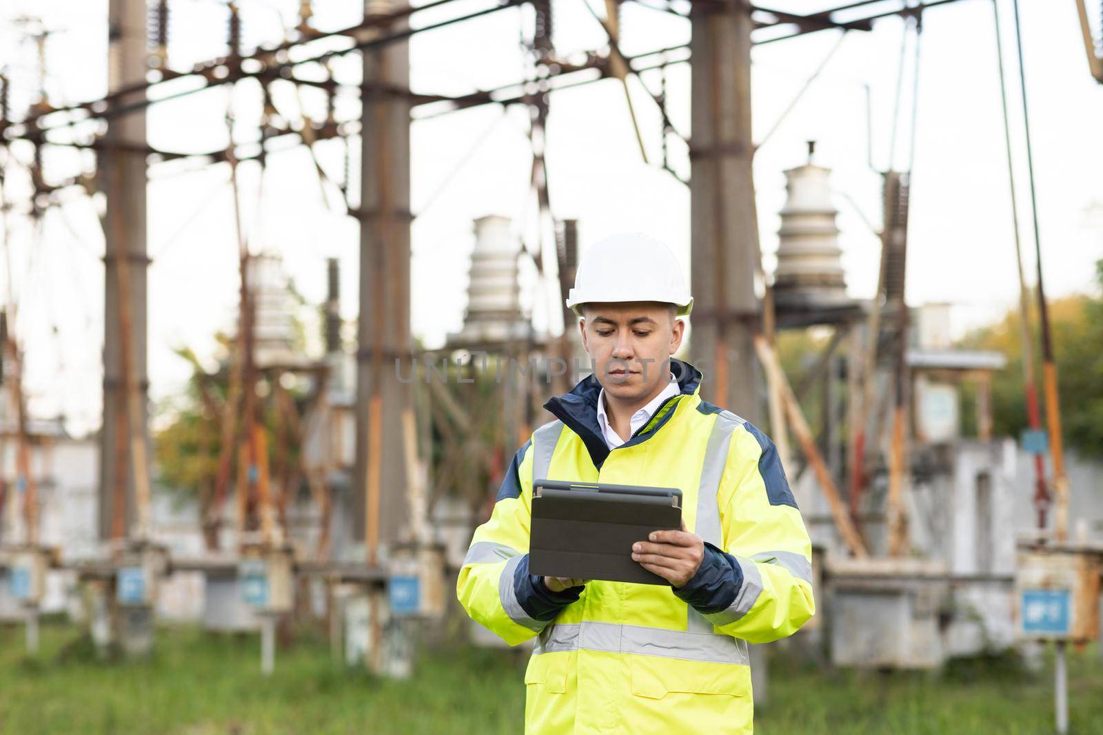 Energy engineer man in special clothes inspects a power line using data from electric sensors on a digital tablet computer. Electrical engineer with high voltage electricity pylon.