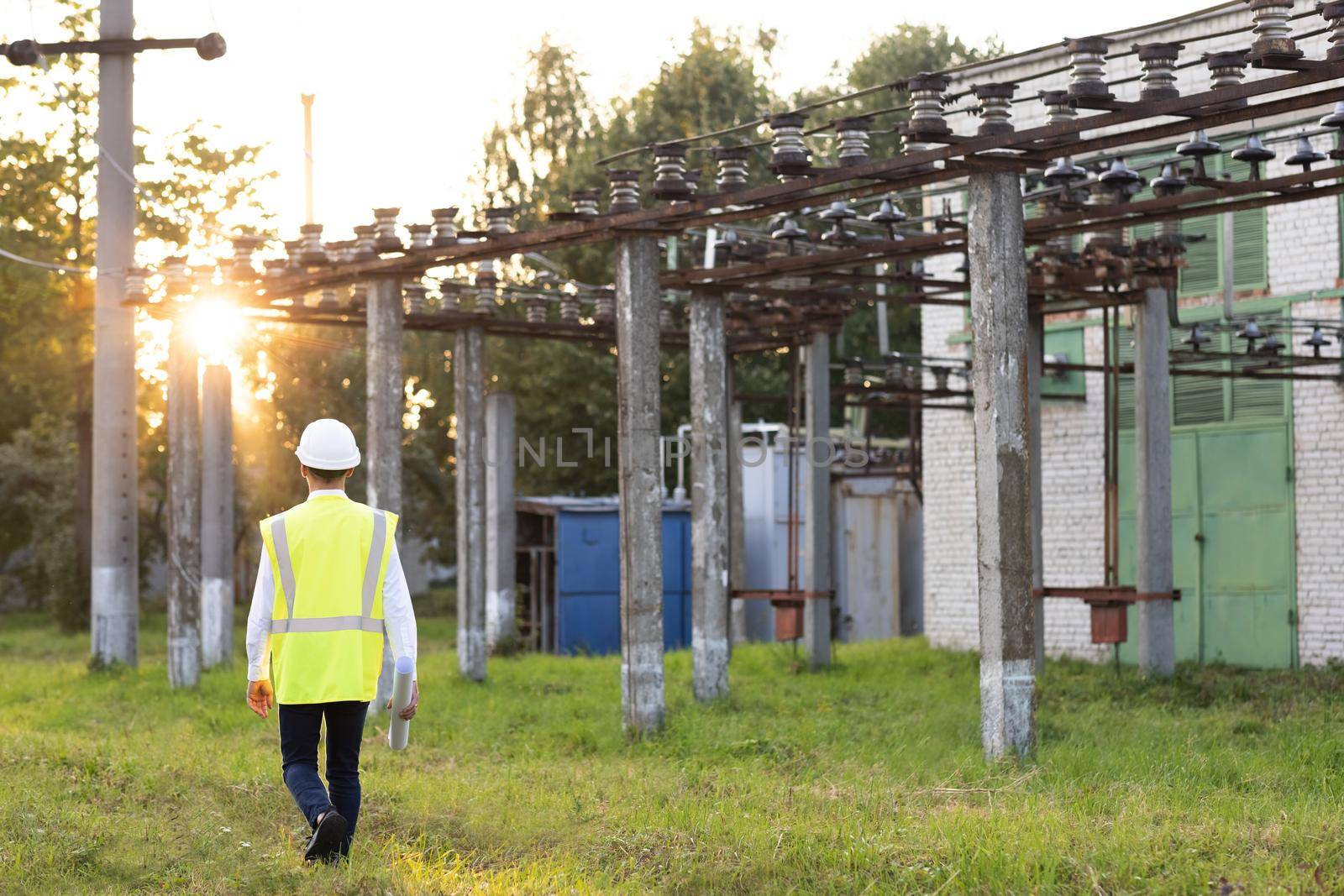 Electrical engineer wearing a helmet and safety vest walking near high voltage electrical lines towards power station during sunset. Concept of electric power station development by uflypro