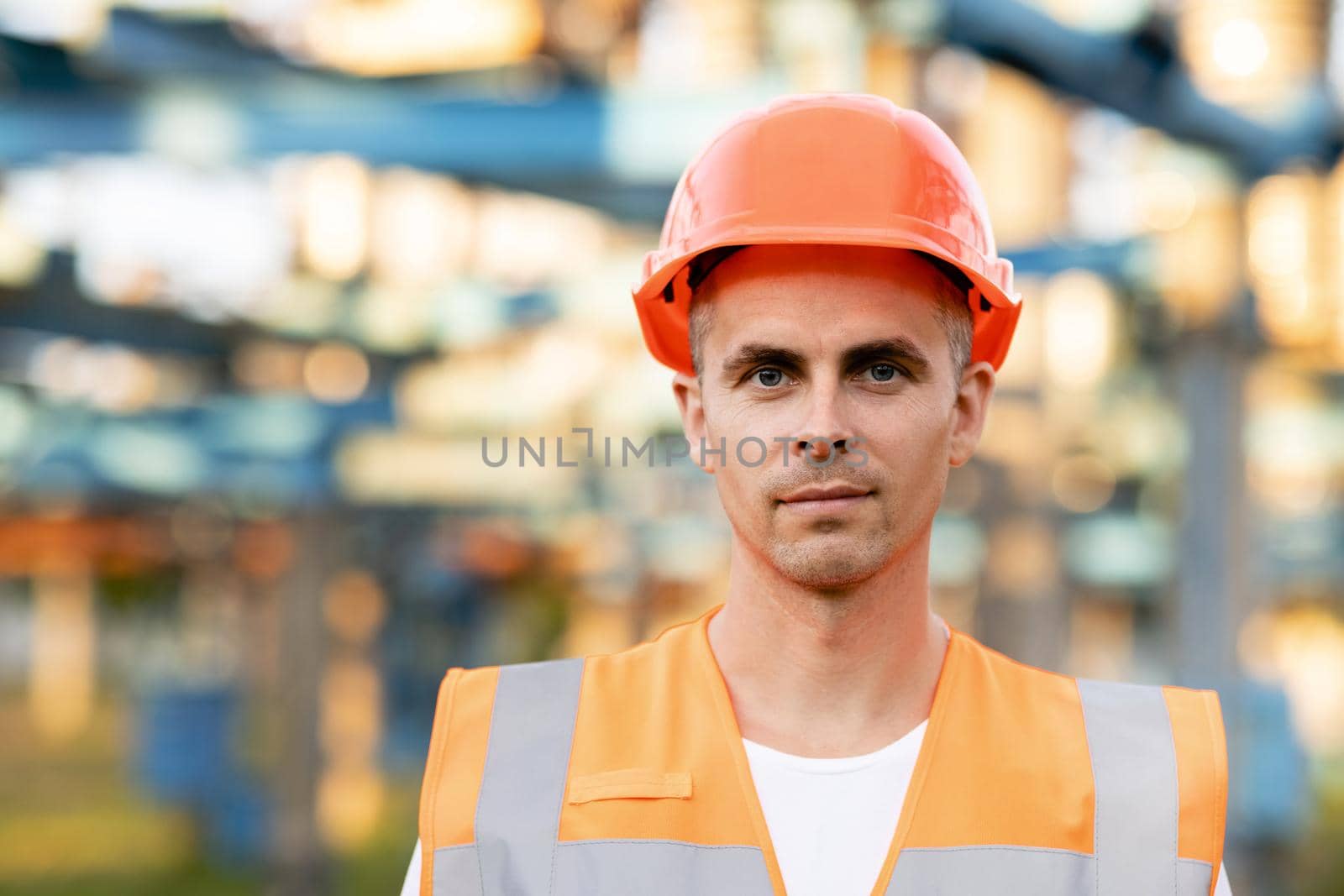 Close up portrait of engineer worker in uniform and helmet standing near high voltage substation with tall pylons and voltage distribution cables by uflypro