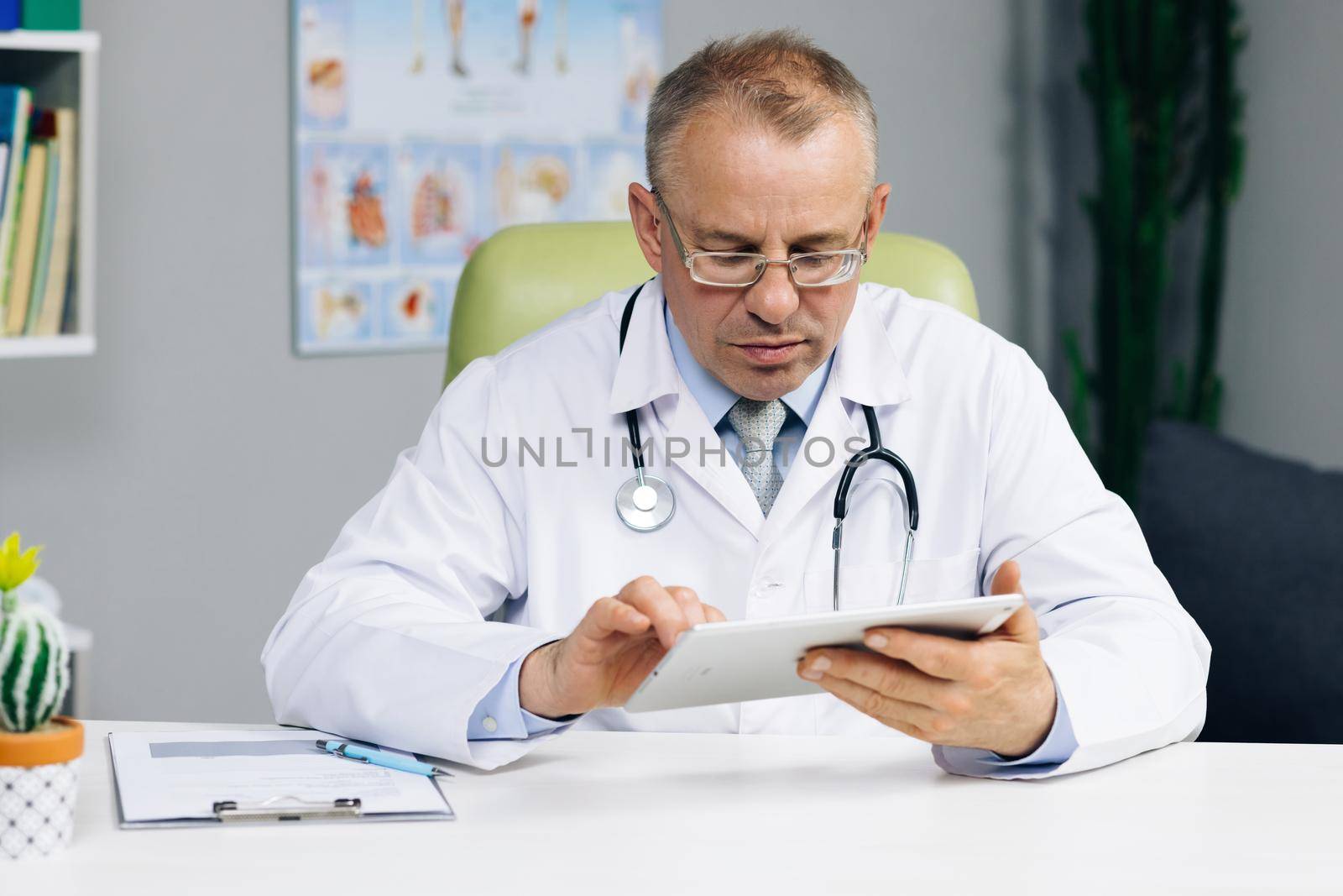 Middle aged older male doctor in white uniform holding digital computer tablet in hands, managing patients visits. Physician checking health history data in gadget, using modern app.