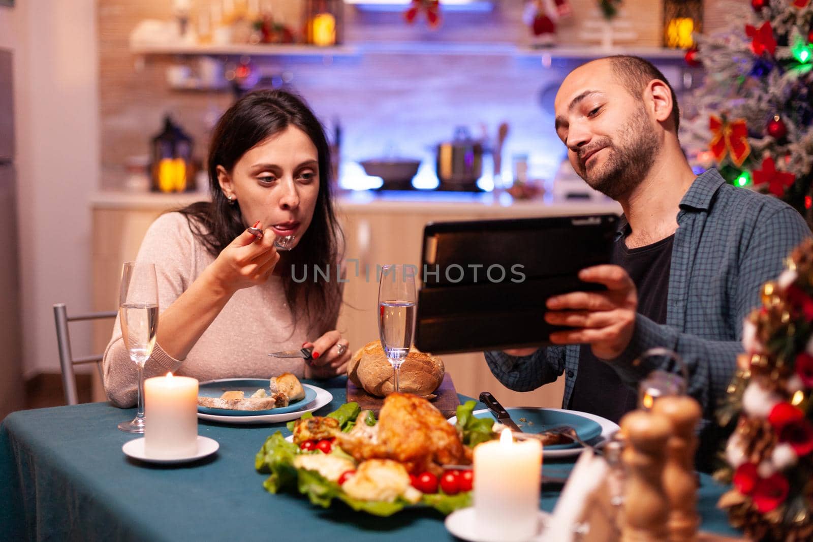 Joyful couple eating delicious dinner sitting at table in xmas decorated kitchen celebrating christmas holiday. Family watching entertainment movie enjoying winter time together. Santa-claus season