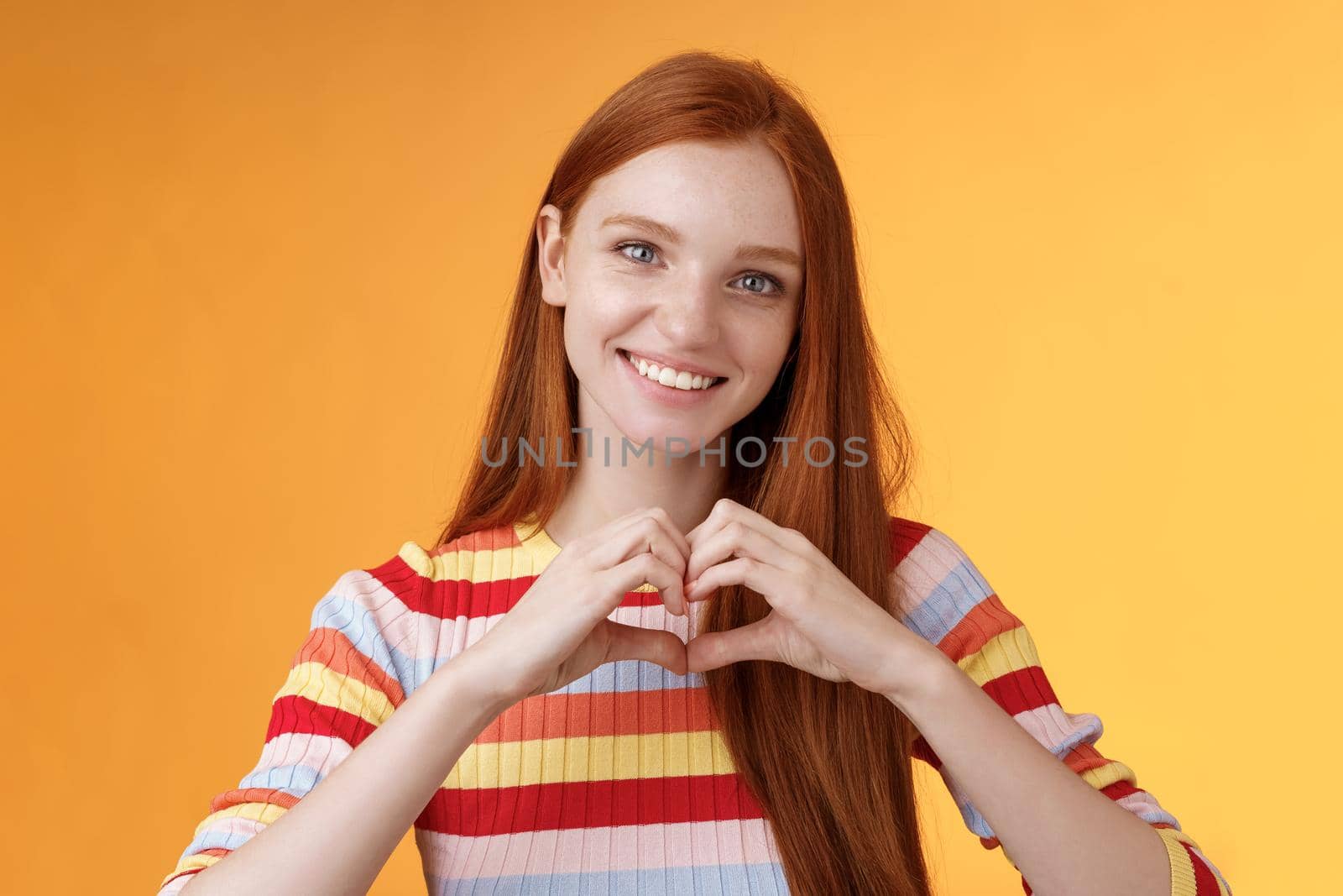 Lovely friendly-looking charismatic smiling redhead girl express affection love and friendship grinning show heart sign chest showing sympathy, confess romantic feelings, standing orange background by Benzoix
