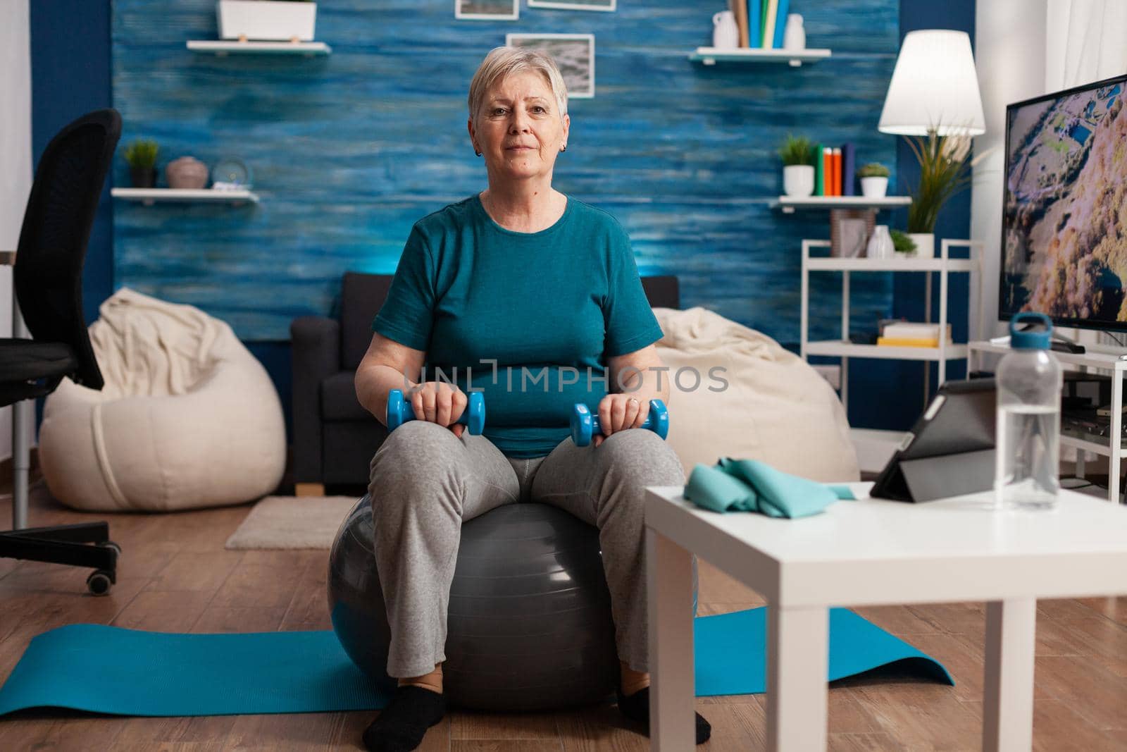 Portrait of senior woman looking into camera while sitting on swiss ball training body muscles holding fitness dumbbells during cardio gym workout. Pensioner working muscular resistance in living room