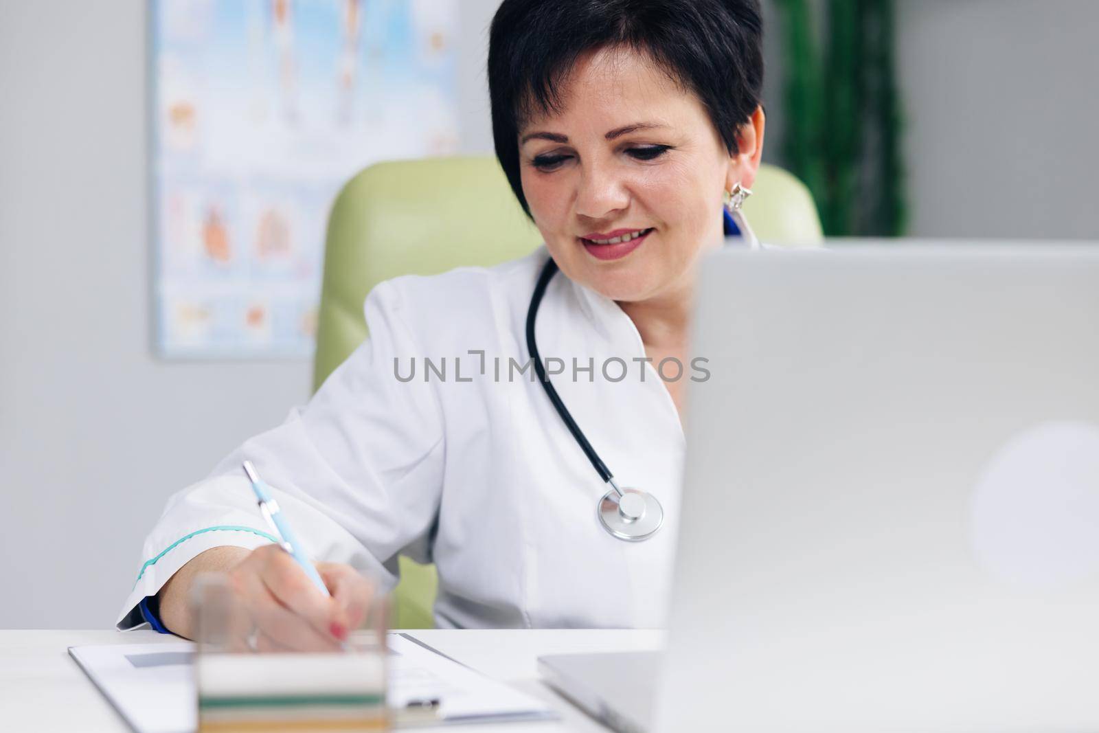 Middle aged senior head doctor in white medical coat sitting at workplace, talking to patient making video call on laptop, writing notes in paper journal.