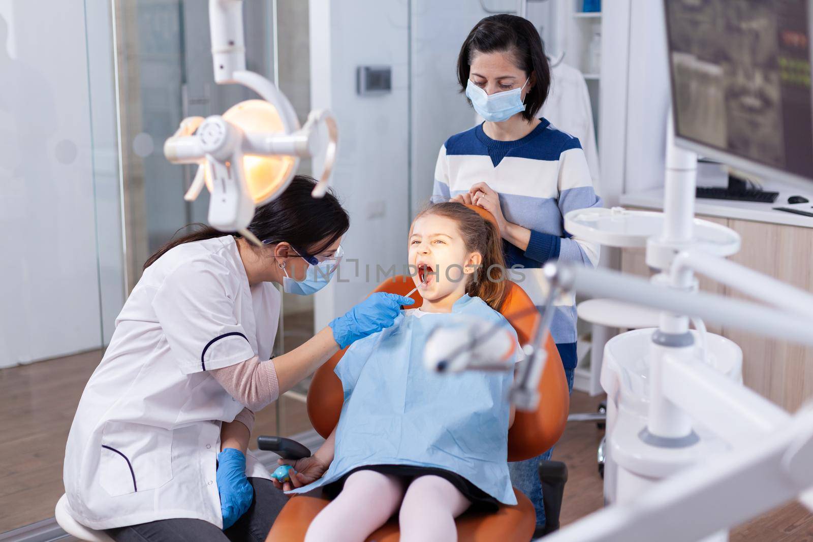 Little girl with open mouth in the course of cavity check up done by stomatologist. Dentistry specialist during child cavity consultation in stomatology office using modern technology.