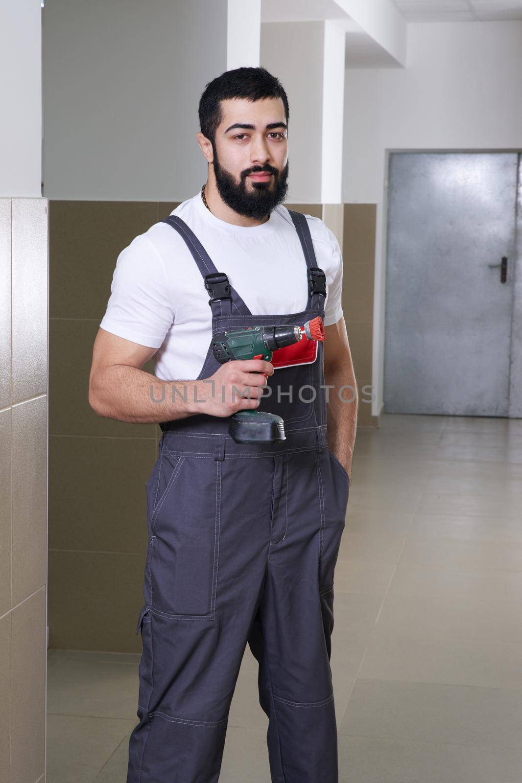 Worker wearing overalls standing and holding electric screwdriver or drill by Mariakray