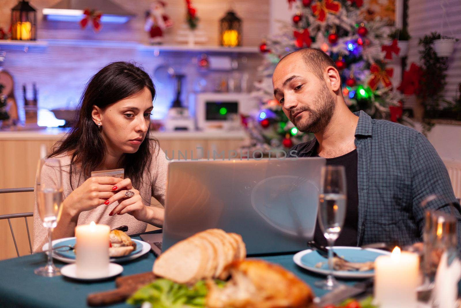 Cheerful couple shopping online xmas gift present paying with credit card on laptop computer sitting at christmas table in x-mas decorated kitchen. Happy family celebrating winter holiday