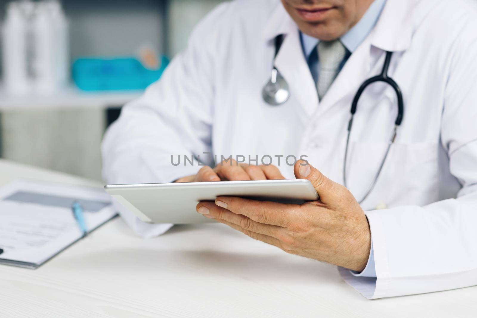 Close up middle aged older male doctor in white uniform holding digital computer tablet in hands, managing patients visits. Physician checking health history data in gadget, using modern app