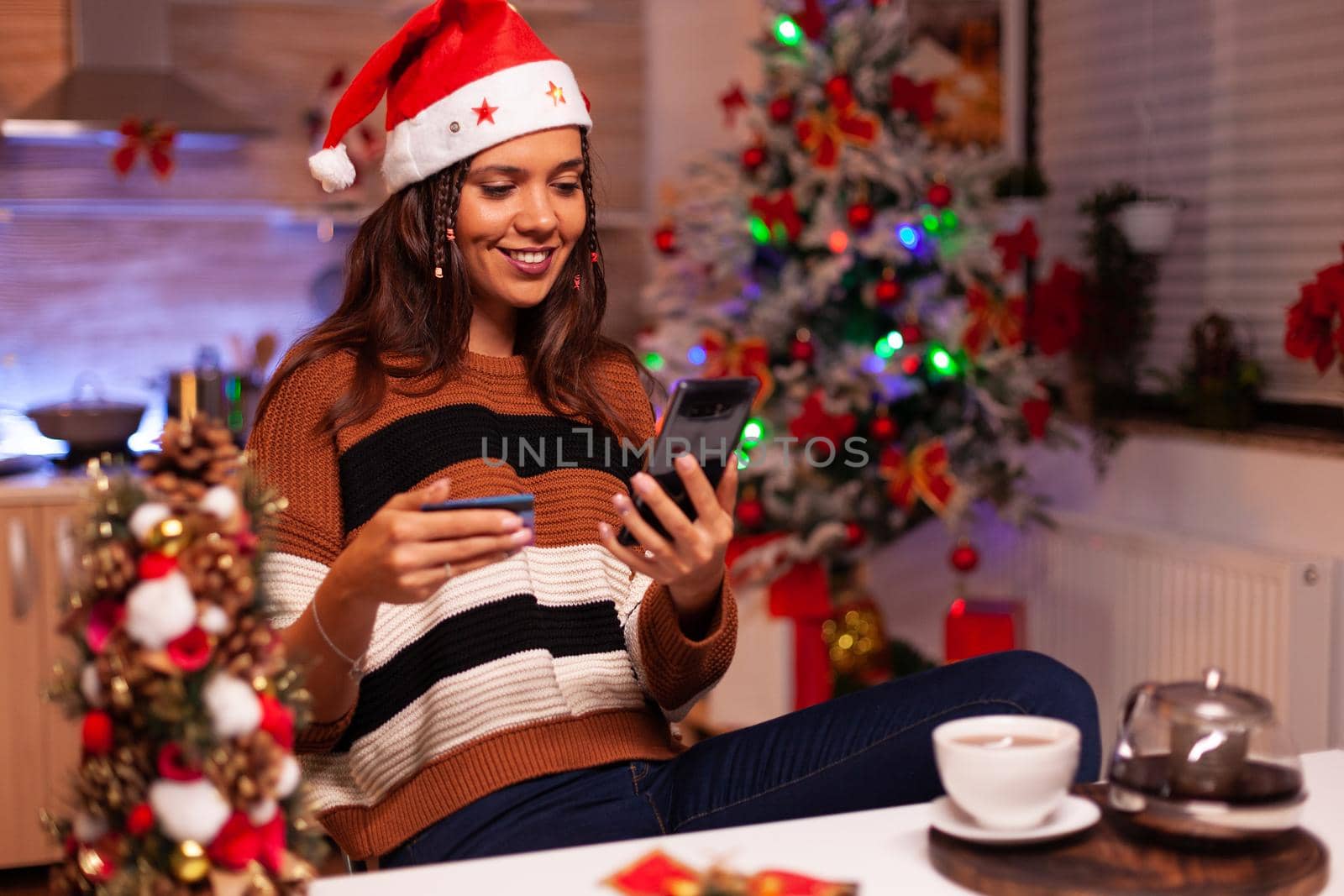 Young woman paying with credit card on shopping site using smartphone at home. Caucasian adult typing number information, ordering christmas gifts online for friends at celebration