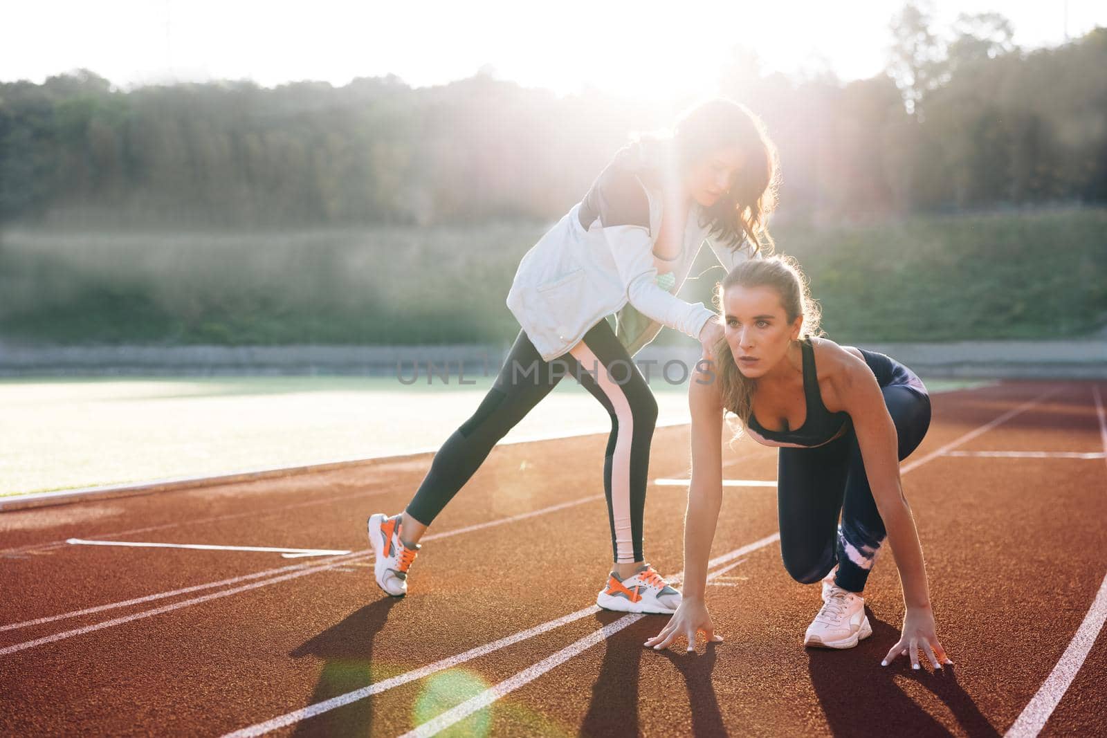 Young female runner with personal trainer preparing for blasting off in mist on sports track of stadium, training before competition. Caucasian Athlete ready to start. Sportswoman. Cardio exercises.