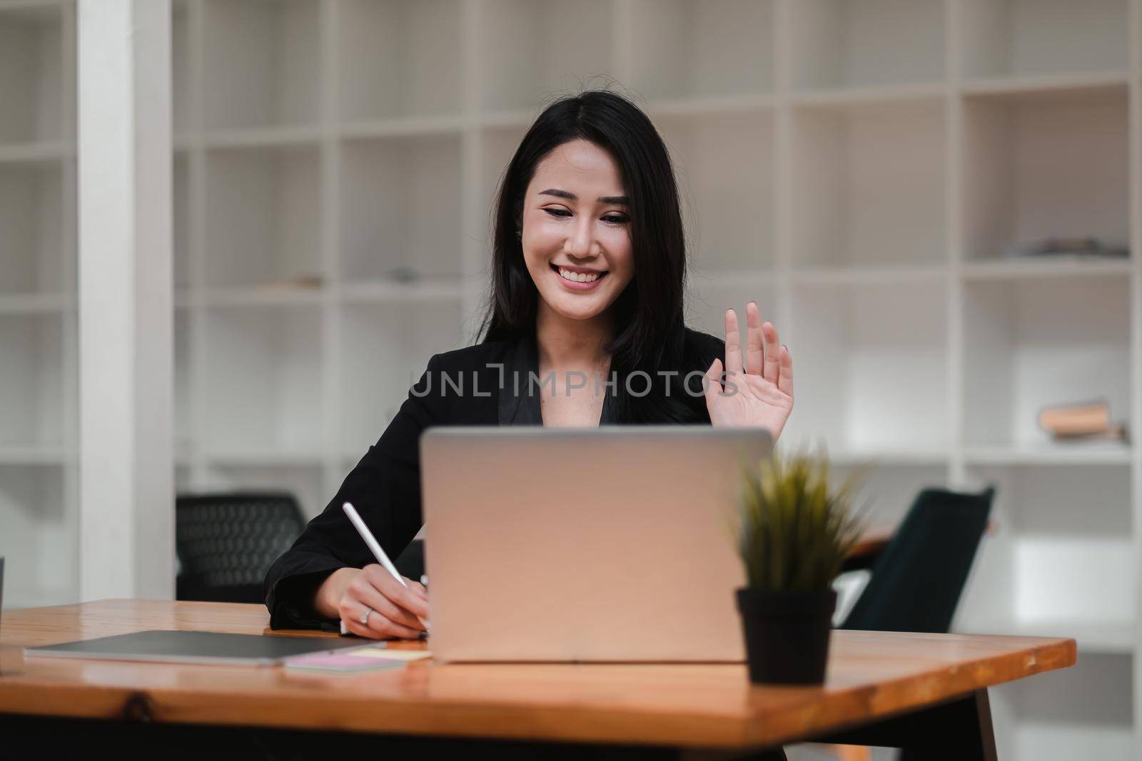 Image of happy asian woman smiling and waving hand at laptop, while speaking or chatting on video call in office by nateemee